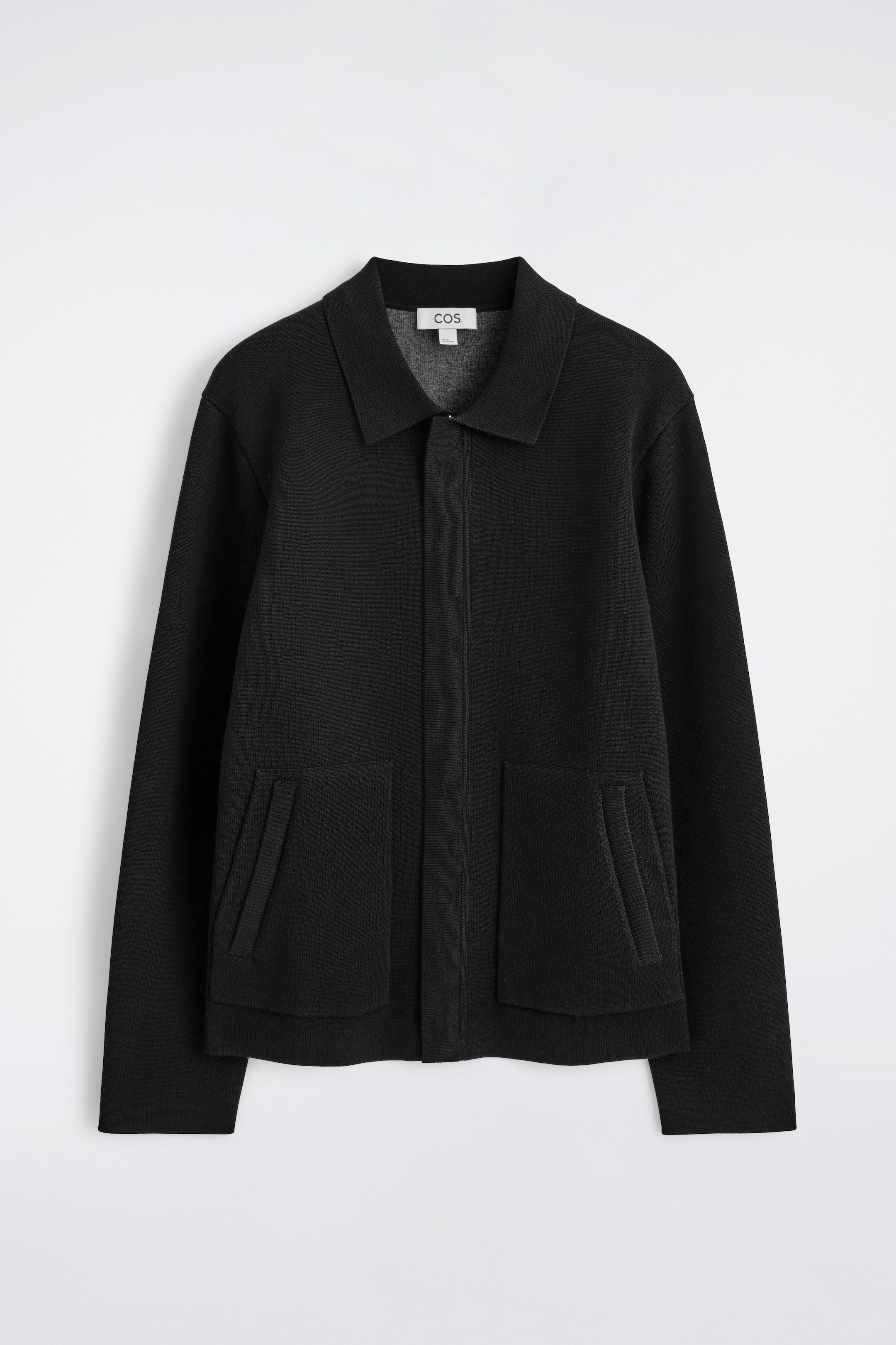 Agrandir l'image: Une veste noire tricotée est affichée de face, avec un col pointu et une fermeture éclair centrale. Elle est conçue avec deux grandes poches plaquées rectangulaires à l'avant, chacune présentant une ouverture en biais.