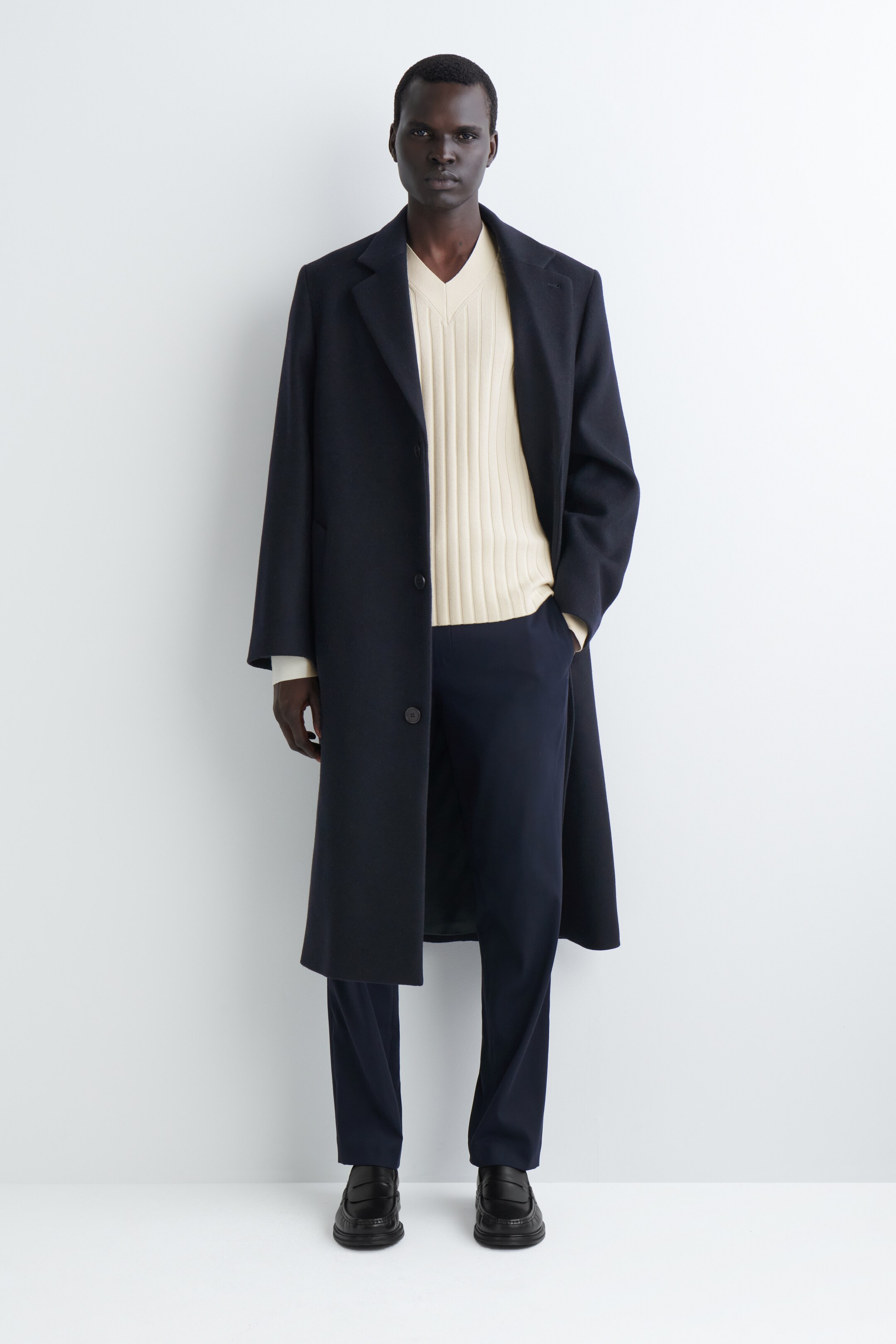 View larger image: A dark, full-length overcoat is being worn, featuring notch lapels and a two-button closure. The coat is being styled open, revealing a light-colored ribbed V-neck sweater and dark trousers underneath, and is draping loosely, extending below the knee.