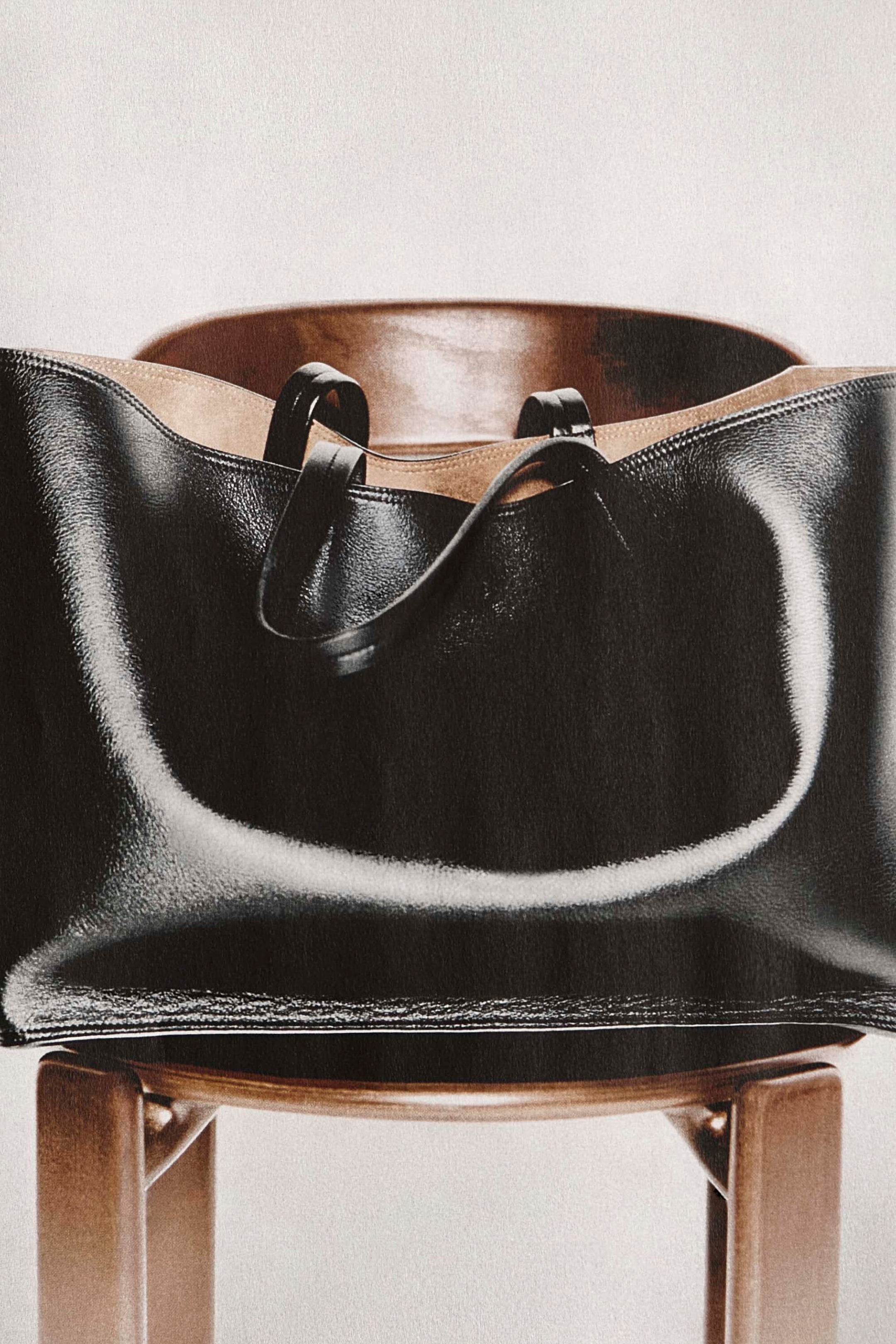 A black textured leather tote bag rests on a wooden chair, highlighting its slouchy, sculpted silhouette from the front. The bag features slim top handles draping inward and a contrasting light tan lining visible along the open top. A black textured leather tote bag rests on a wooden chair, highlighting its slouchy, sculpted silhouette from the front. The bag features slim top handles draping inward and a contrasting light tan lining visible along the open top.