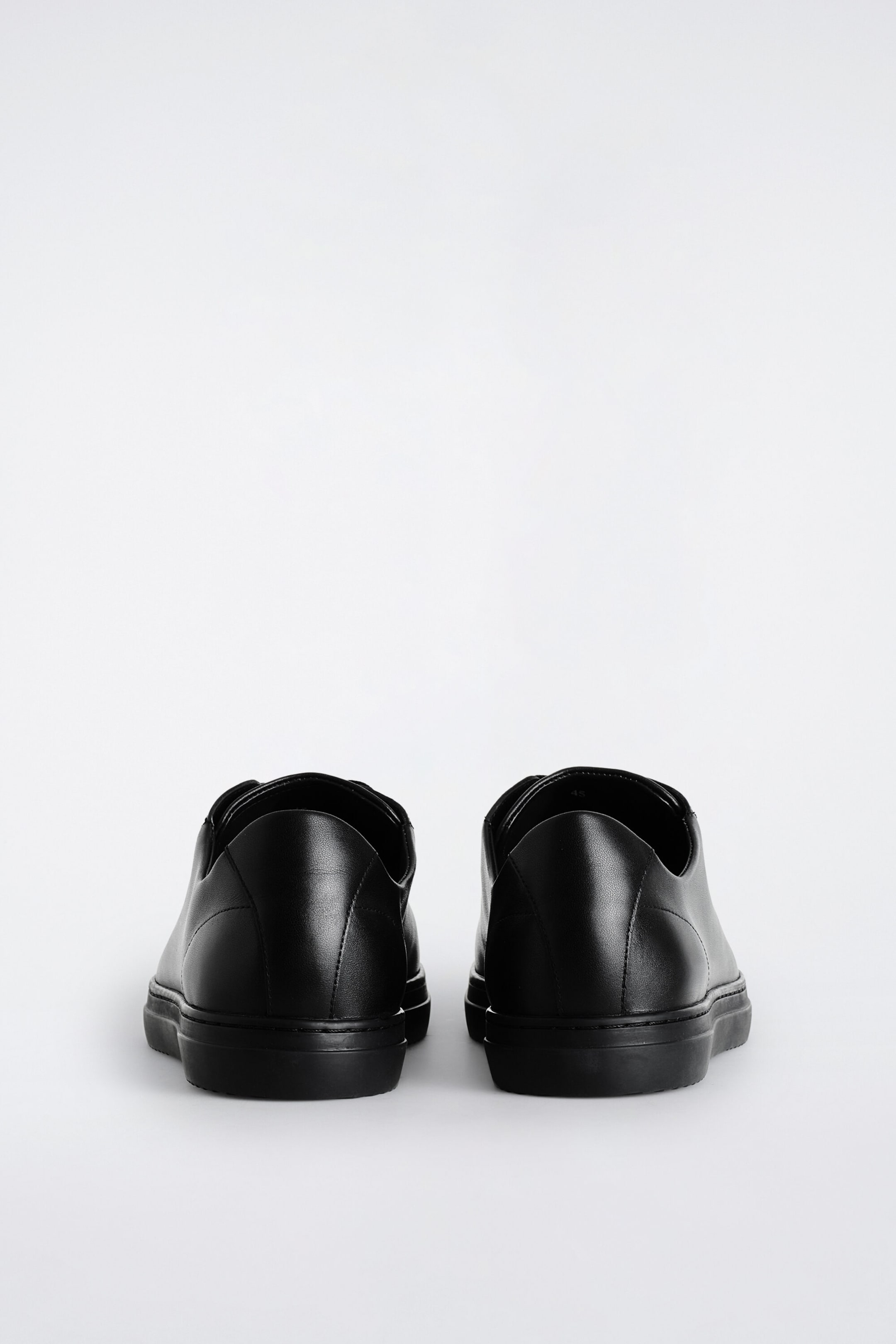Leather sneakers are positioned facing away, showcasing their minimalist design in smooth black leather. They feature rounded heel counters with clean tonal stitching and matching black rubber soles. Leather sneakers are positioned facing away, showcasing their minimalist design in smooth black leather. They feature rounded heel counters with clean tonal stitching and matching black rubber soles.
