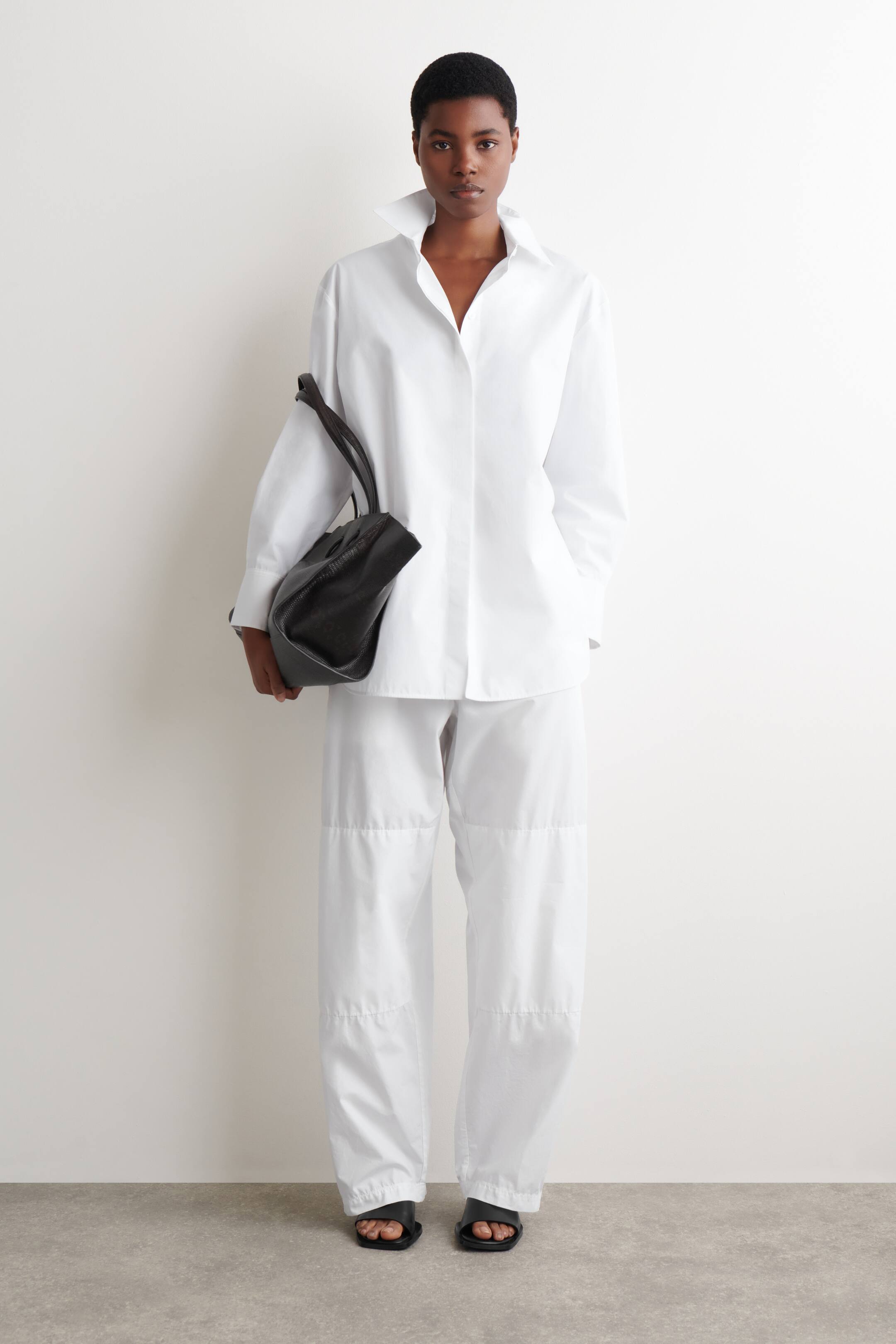 Agrandir l'image: Une chemise blanche oversize, présentant un col montant et des manches longues, est portée avec un pantalon blanc ample comportant des panneaux de couture horizontaux. Un sac d'épaule noir non structuré est tenu et des sandales plates noires à enfiler sont portées.