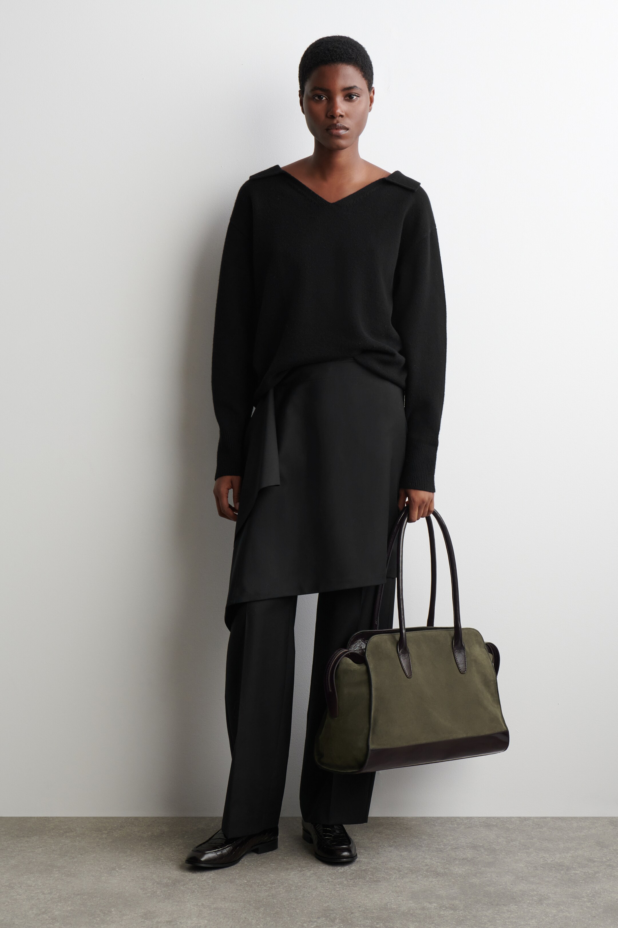 View larger image: A black V-neck sweater featuring wide, folded shoulder details and long sleeves is being worn over a black asymmetrical wrap overlay and black wide-leg trousers. The ensemble is completed with black leather shoes and an olive green canvas and dark brown leather tote bag, being held by its top handles and facing slightly forward.
