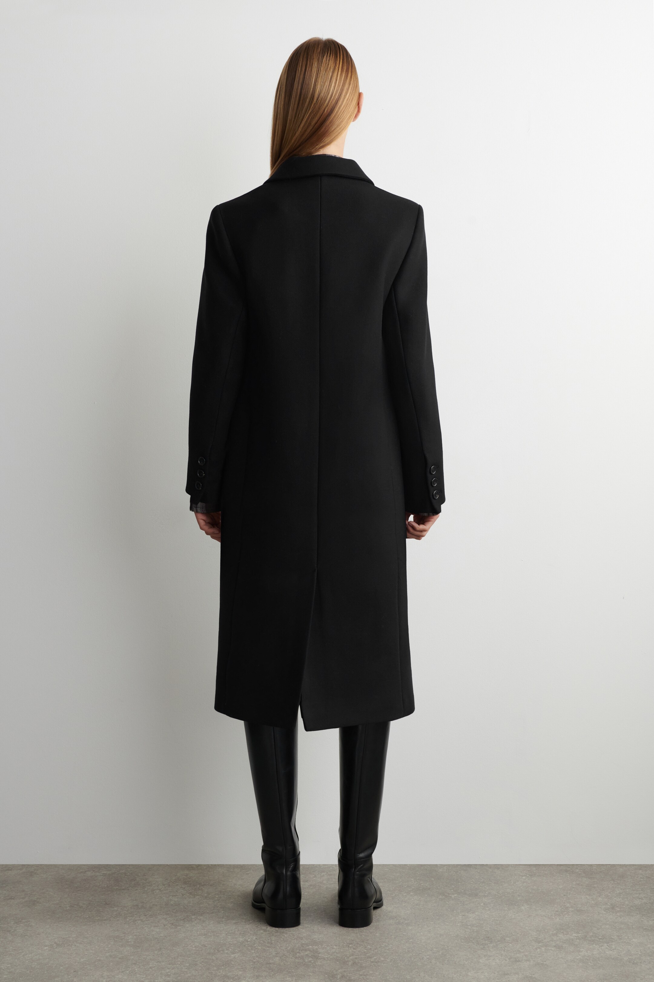 Ingrandisci l'immagine: Un lungo cappotto nero, con uno spacco posteriore e dettagli di bottoni sui polsini, è indossato dalla modella. Il cappotto arriva ai polpacci della modella, rivelando alti stivali neri.