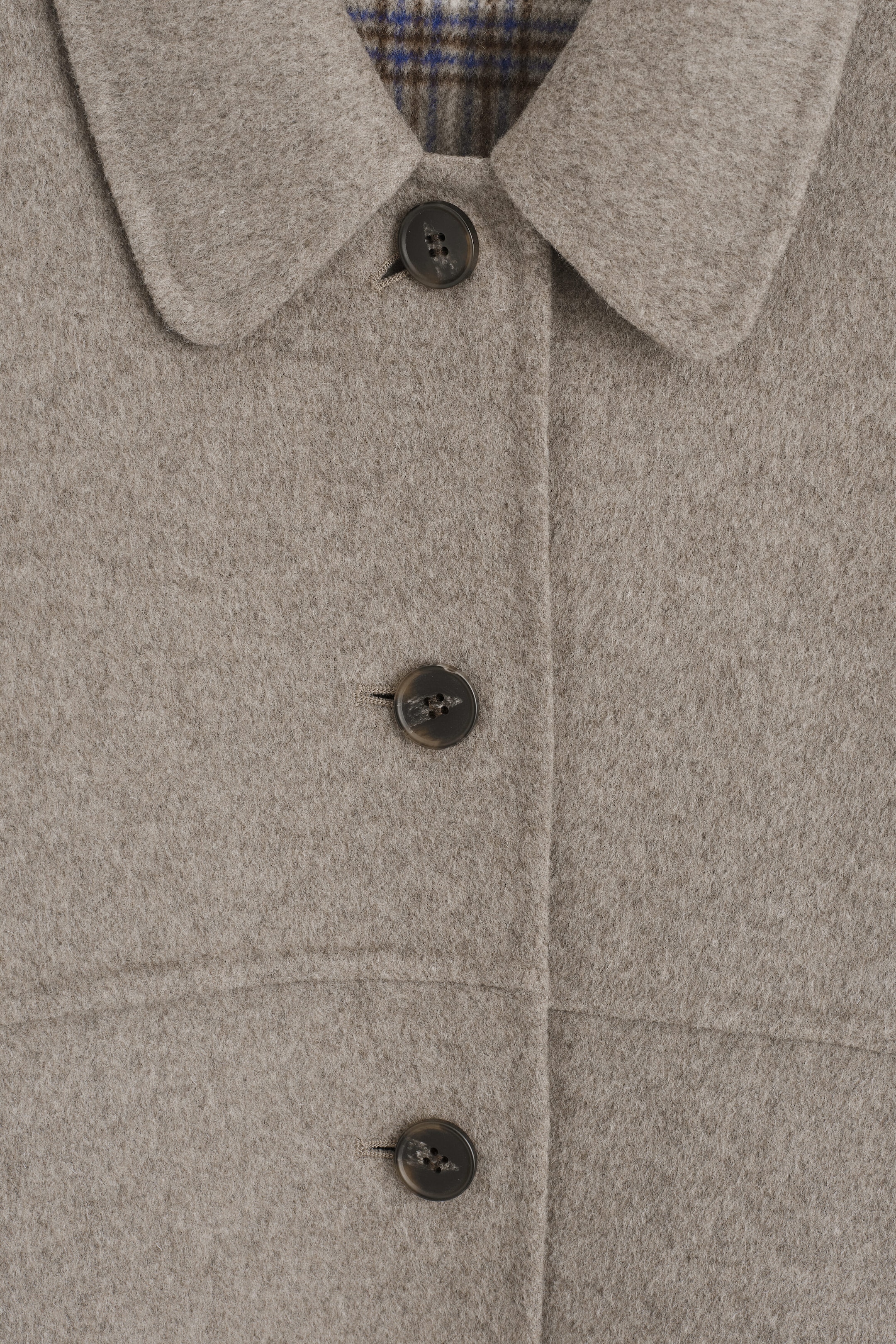 A close-up front view of a beige wool coat, showing its soft, brushed texture and featuring a classic point collar with dark button closures. A glimpse of the blue and brown checkered lining is visible at the inner neck, showcasing the coat's reversible design.