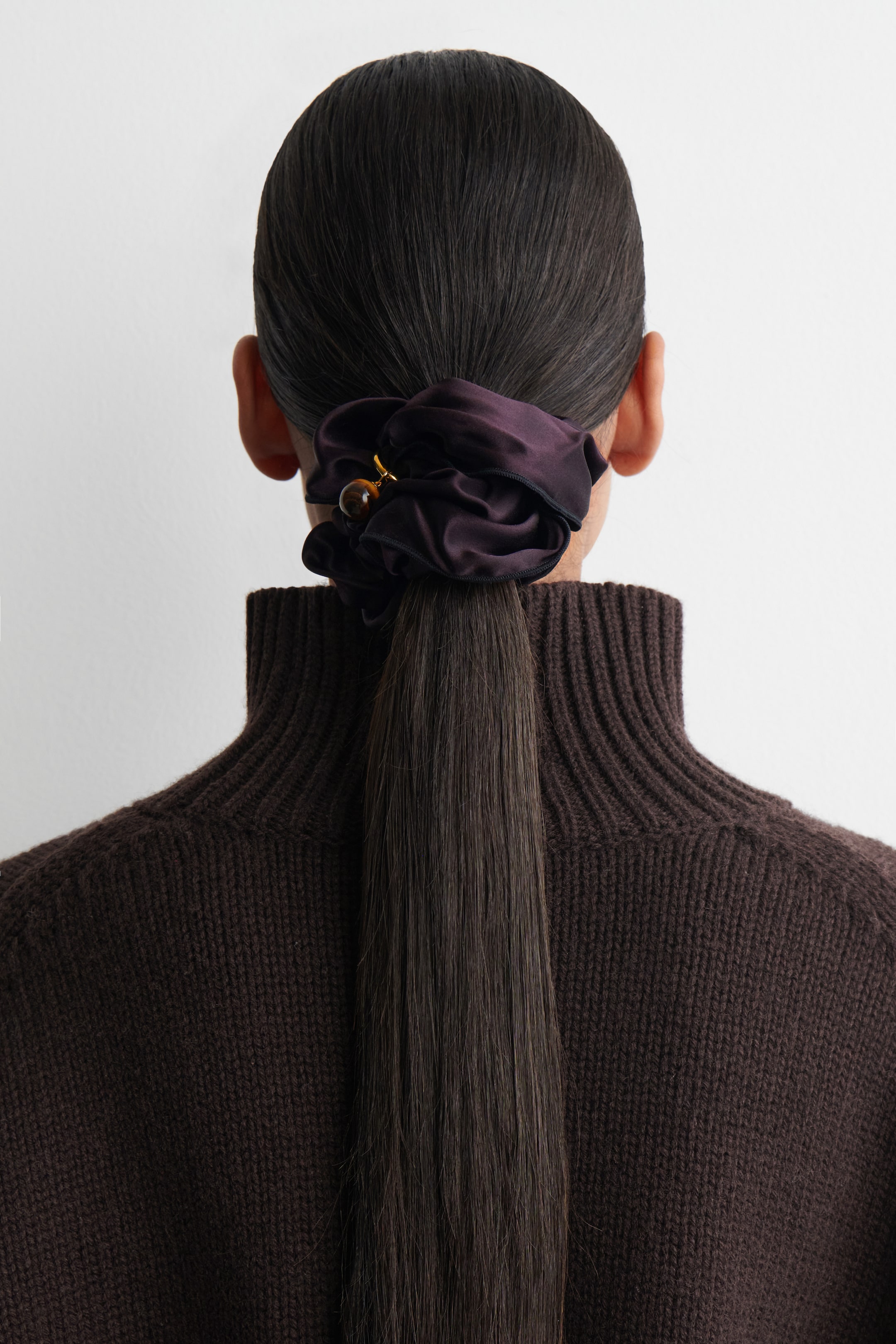 Visualizza immagine più grande: Un elastico per capelli in raso color prugna scuro sta fissando una coda bassa, vista da dietro. Lo scrunchie appare voluminoso e raccolto, caratterizzato da un piccolo ciondolo dorato con una perla sferica marrone marmorizzata.