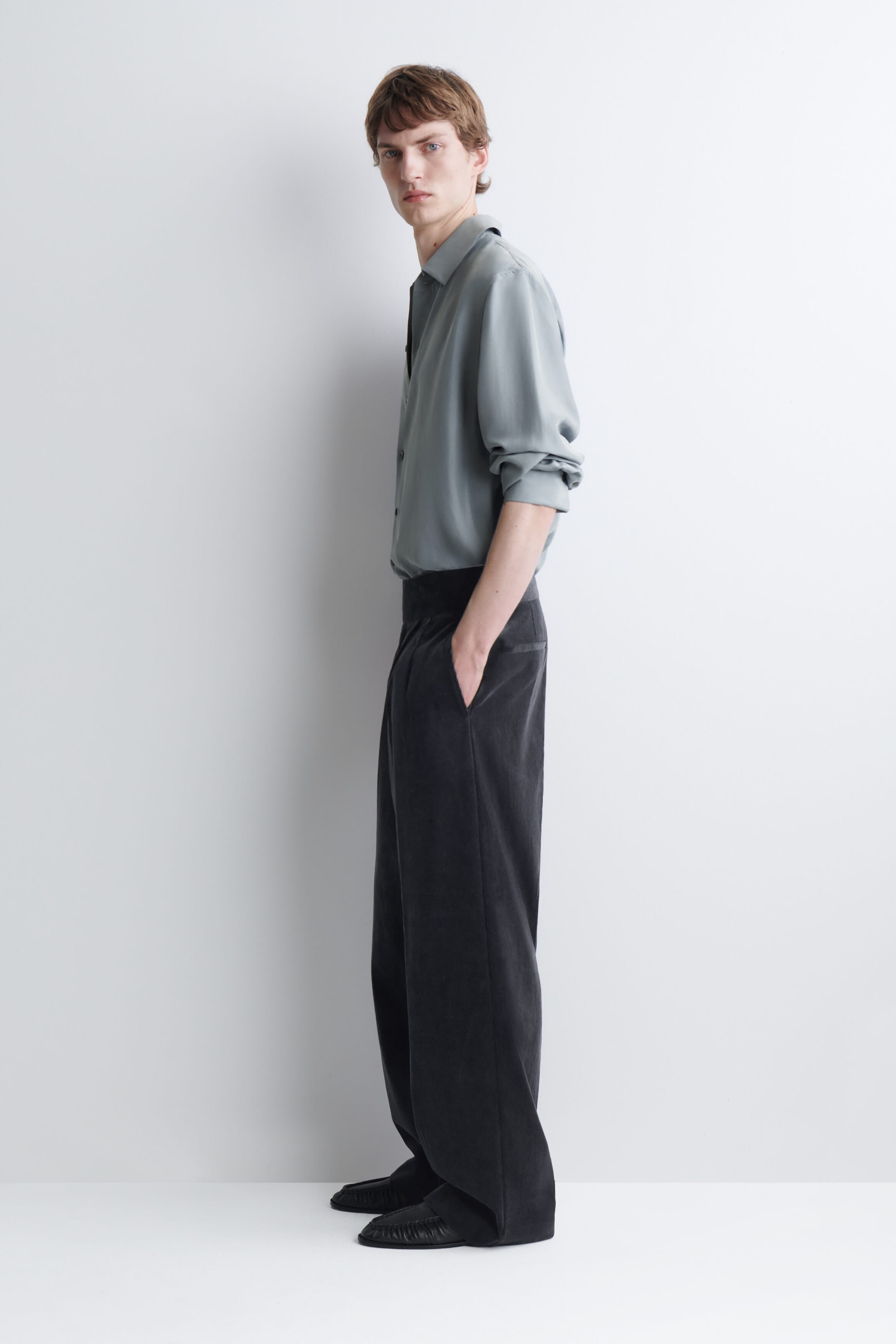 The model is wearing dark gray, relaxed-fit corduroy wide-leg pants and standing in a side-profile pose with one hand resting in the side pocket. The high-waisted pants are styled with a tucked-in light gray-green long-sleeve shirt and black leather shoes. The model is wearing dark gray, relaxed-fit corduroy wide-leg pants and standing in a side-profile pose with one hand resting in the side pocket. The high-waisted pants are styled with a tucked-in light gray-green long-sleeve shirt and black leather shoes.