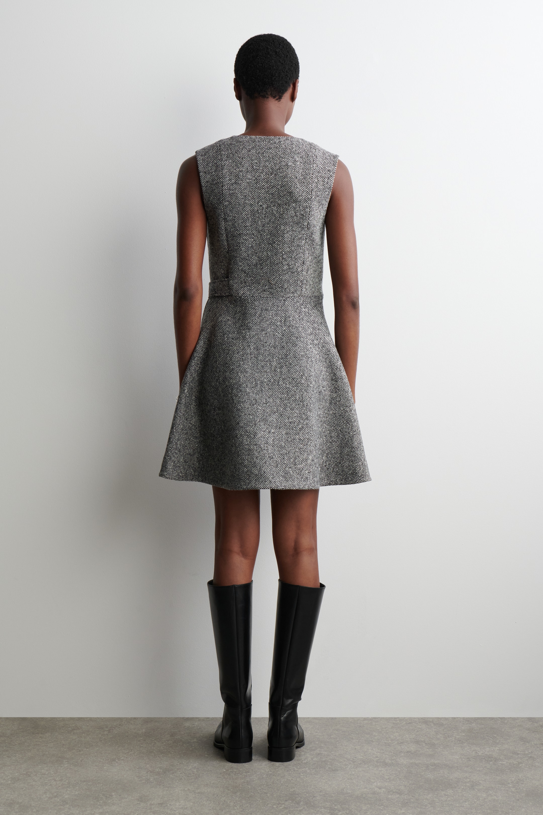 Ingrandisci l'immagine: Un vestito grigio senza maniche con effetto screziato è presentato dal retro, caratterizzato da un corpetto aderente, una vita definita e una gonna svasata a linea A che arriva sopra il ginocchio. Stivali neri al ginocchio vengono indossati con l'abito.