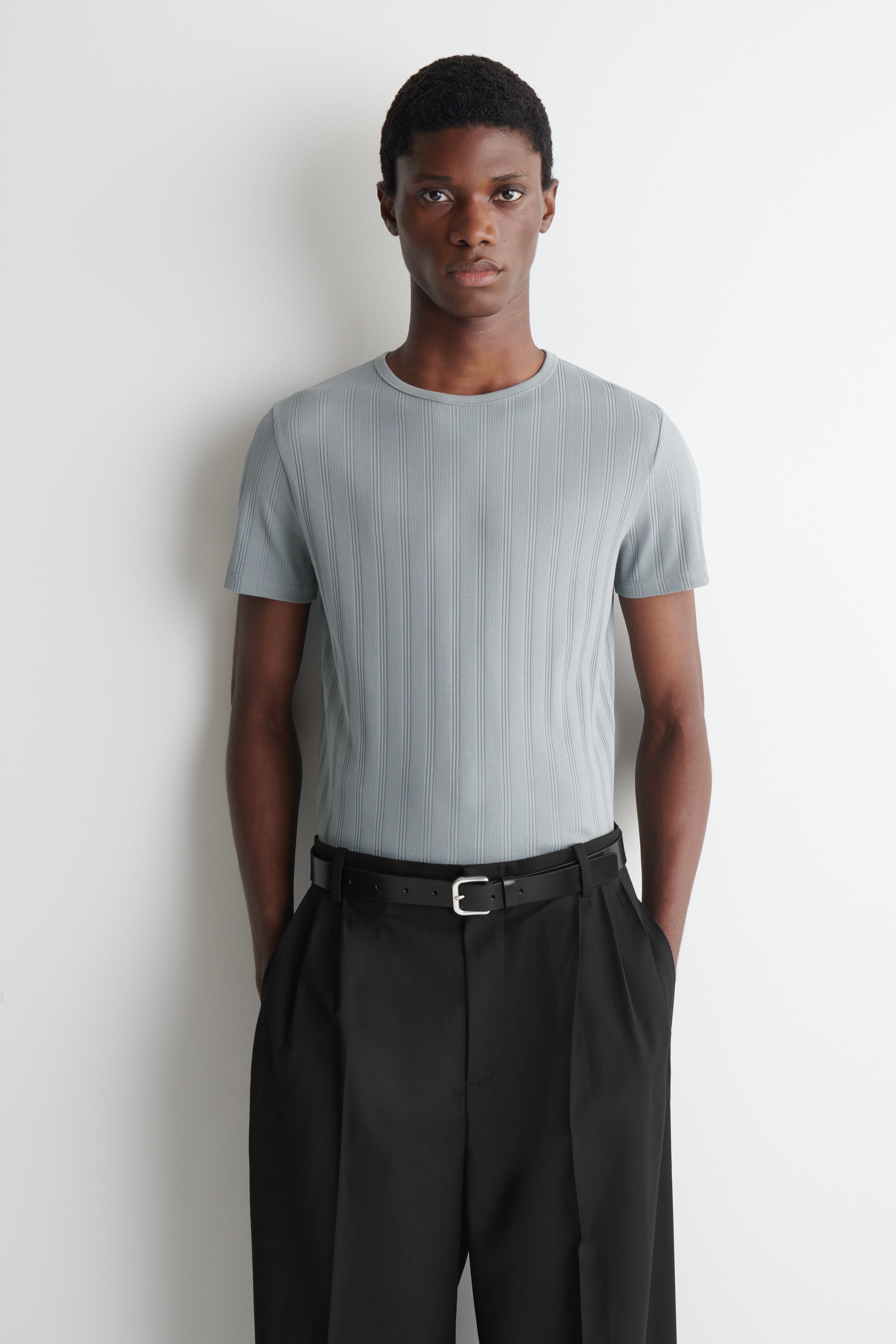 Una t-shirt grigio pietra è presentata frontalmente. Presenta uno scollo a girocollo, maniche corte e una texture a coste larghe verticali che si estende su tutta la superficie.