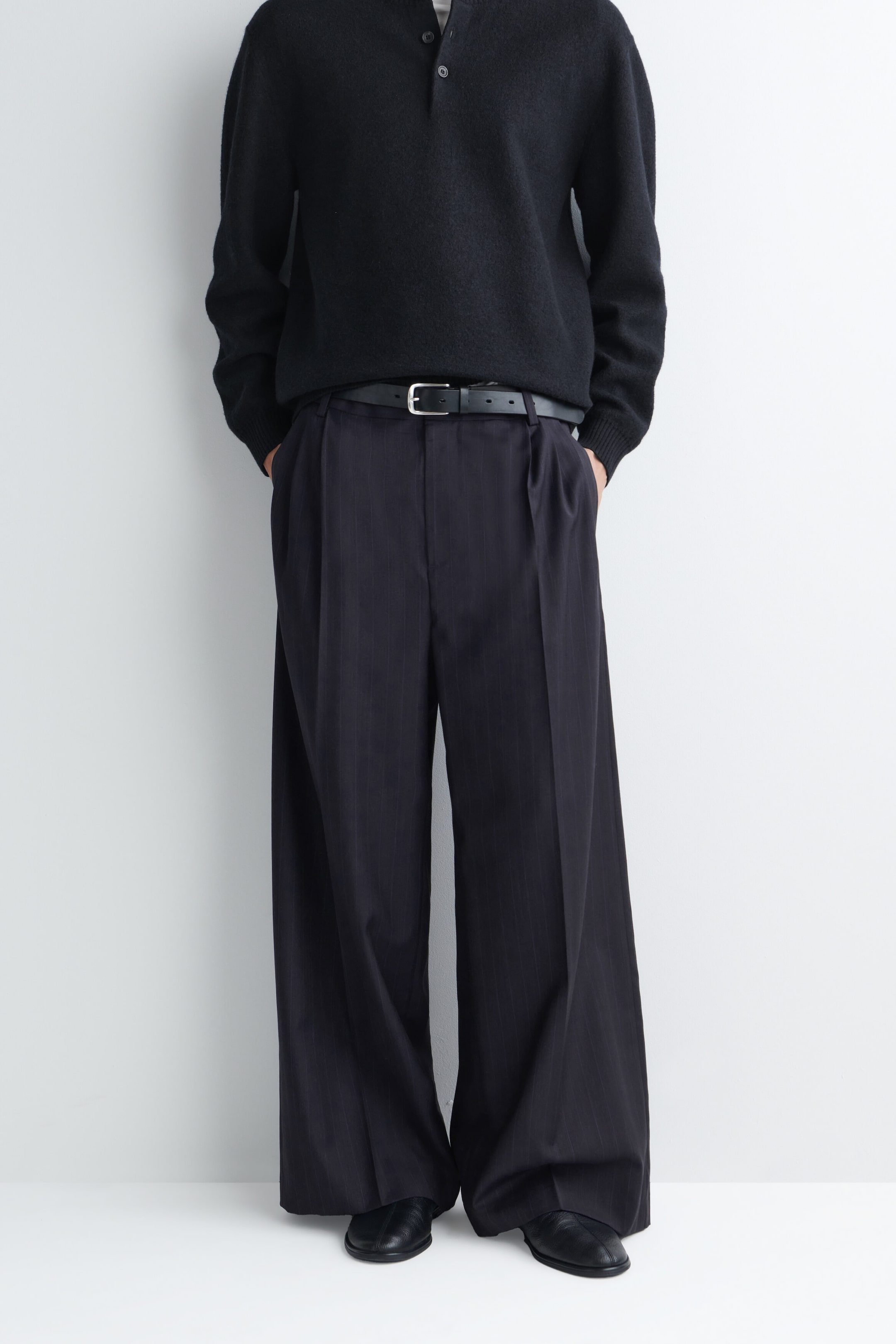 Pantalon à jambe large à coupe décontractée avec plis, en tissu sombre à fines rayures, porté par le modèle, qui se tient debout les mains posées dans les poches latérales. Le pantalon présente des plis à l’avant et une silhouette ample, agencée avec une ceinture en cuir noir, un chandail en tricot noir et des chaussures en cuir noir. Pantalon à jambe large à coupe décontractée avec plis, en tissu sombre à fines rayures, porté par le modèle, qui se tient debout les mains posées dans les poches latérales. Le pantalon présente des plis à l’avant et une silhouette ample, agencée avec une ceinture en cuir noir, un chandail en tricot noir et des chaussures en cuir noir.
