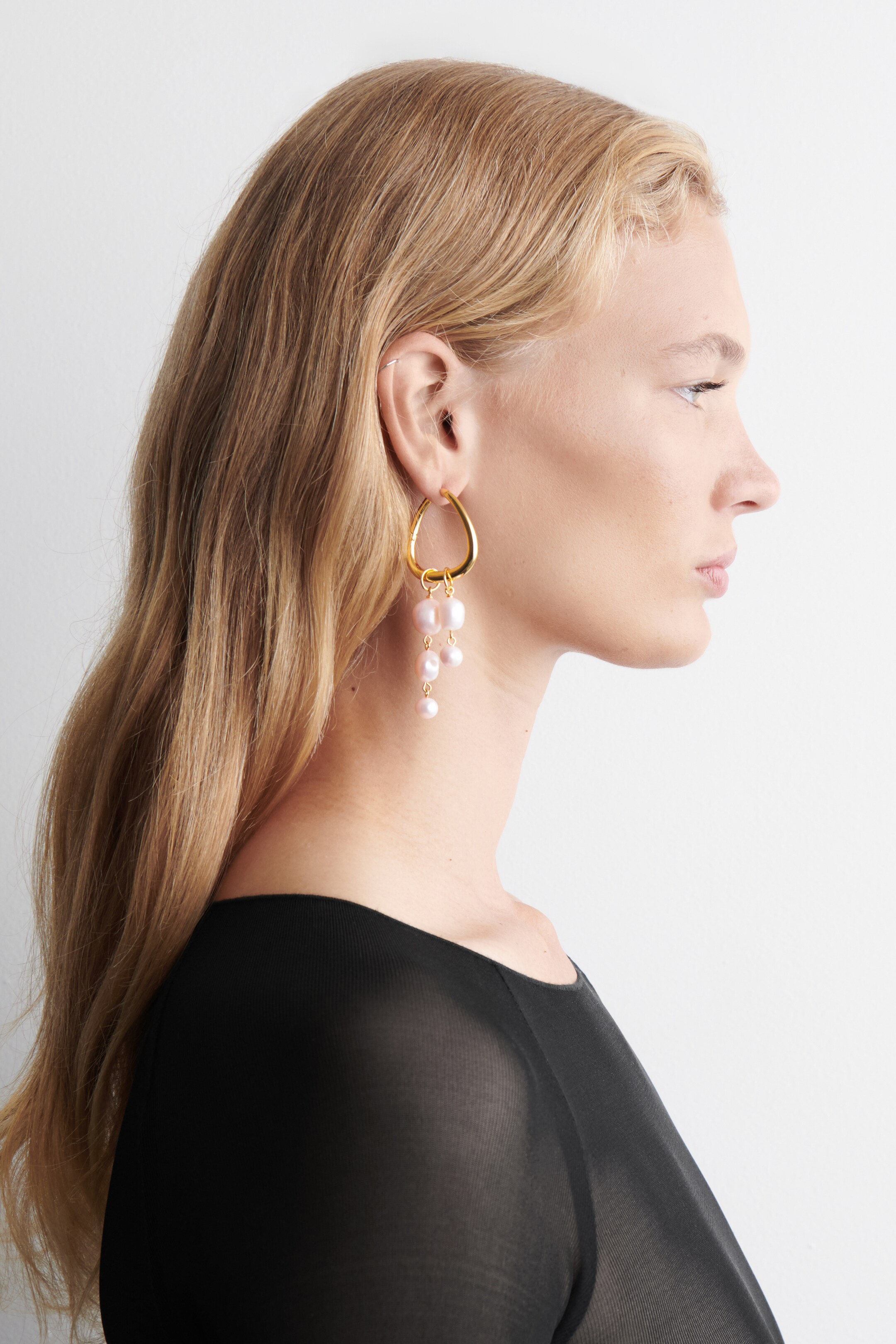 View larger image: A gold-toned, tear-drop shaped hoop earring is hanging from the right ear, viewed from a right-side profile. Cascading clusters of light pink pearls are dangling from the base of the hoop, varying in size.