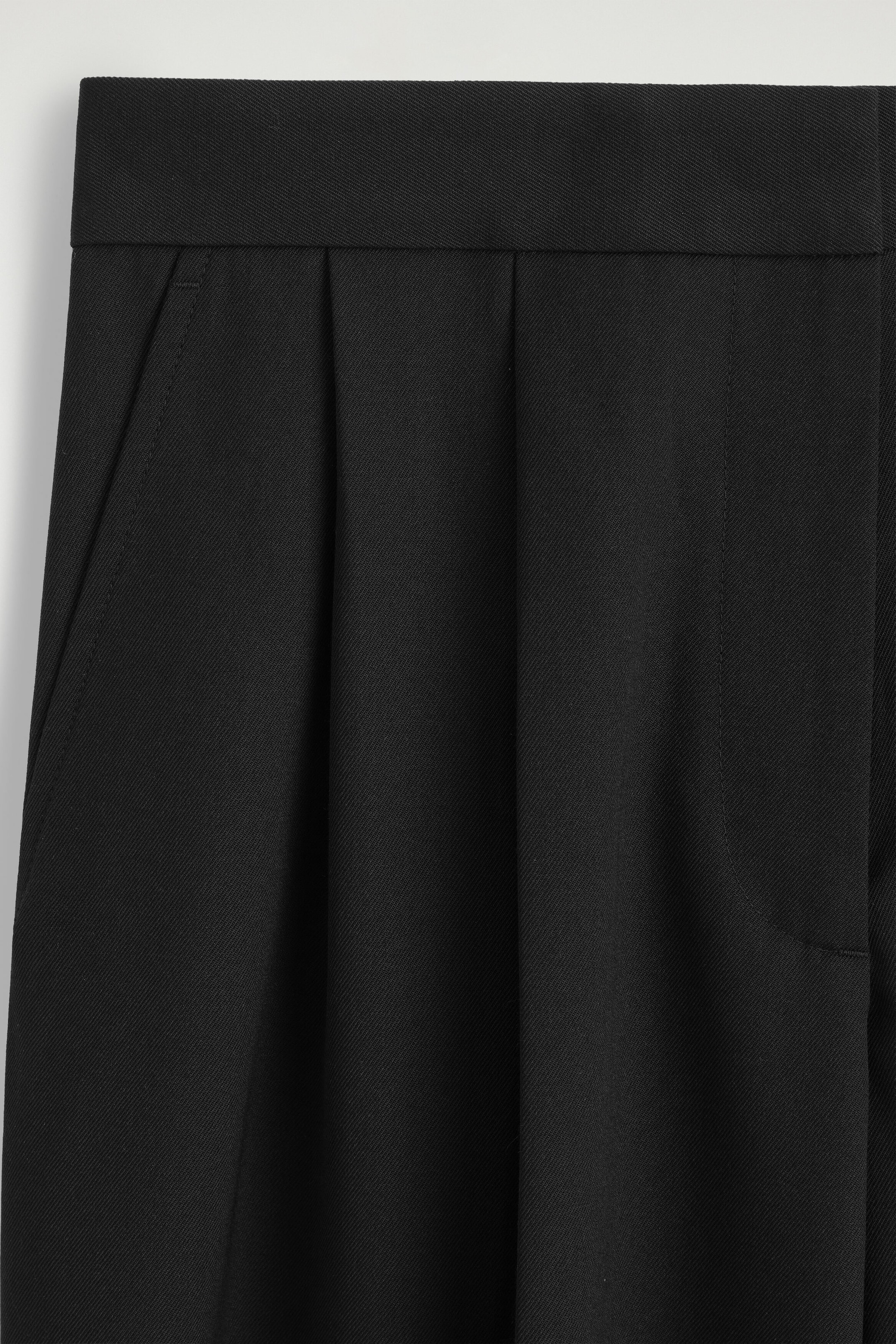 The black pleated wide-leg pants are shown in a close-up front view, highlighting a structured waistband and deep front pleats. The diagonal twill fabric drapes downward, with a side slip pocket visible on the left. The black pleated wide-leg pants are shown in a close-up front view, highlighting a structured waistband and deep front pleats. The diagonal twill fabric drapes downward, with a side slip pocket visible on the left.