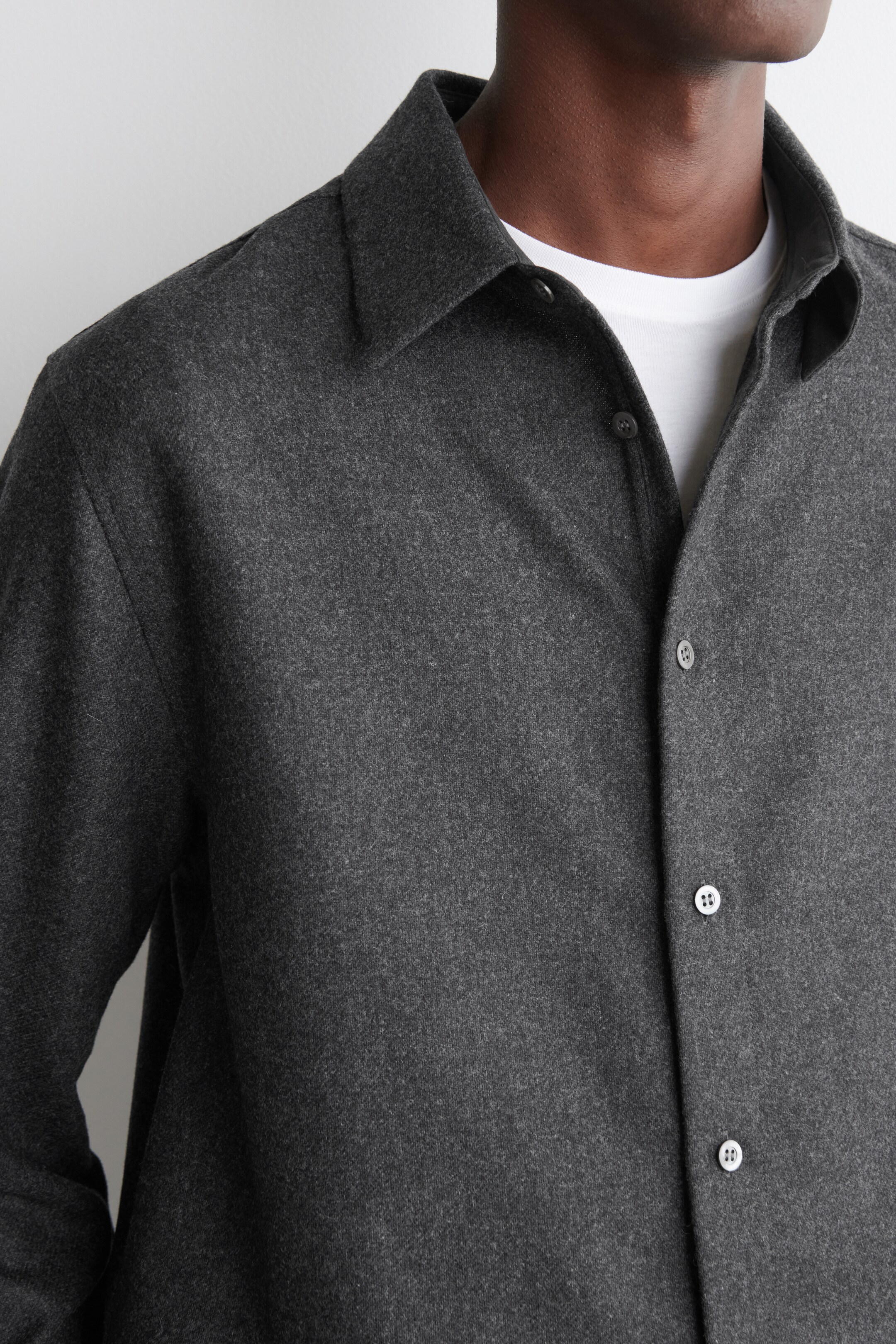 Visualizza immagine più grande: Una camicia grigio scuro con una superficie spazzolata viene presentata da un'angolazione frontale-leggermente a destra. Viene indossata aperta sopra una t-shirt bianca con scollo rotondo, caratterizzata da un colletto a punta e bottoni di colore chiaro lungo l'abbottonatura frontale.