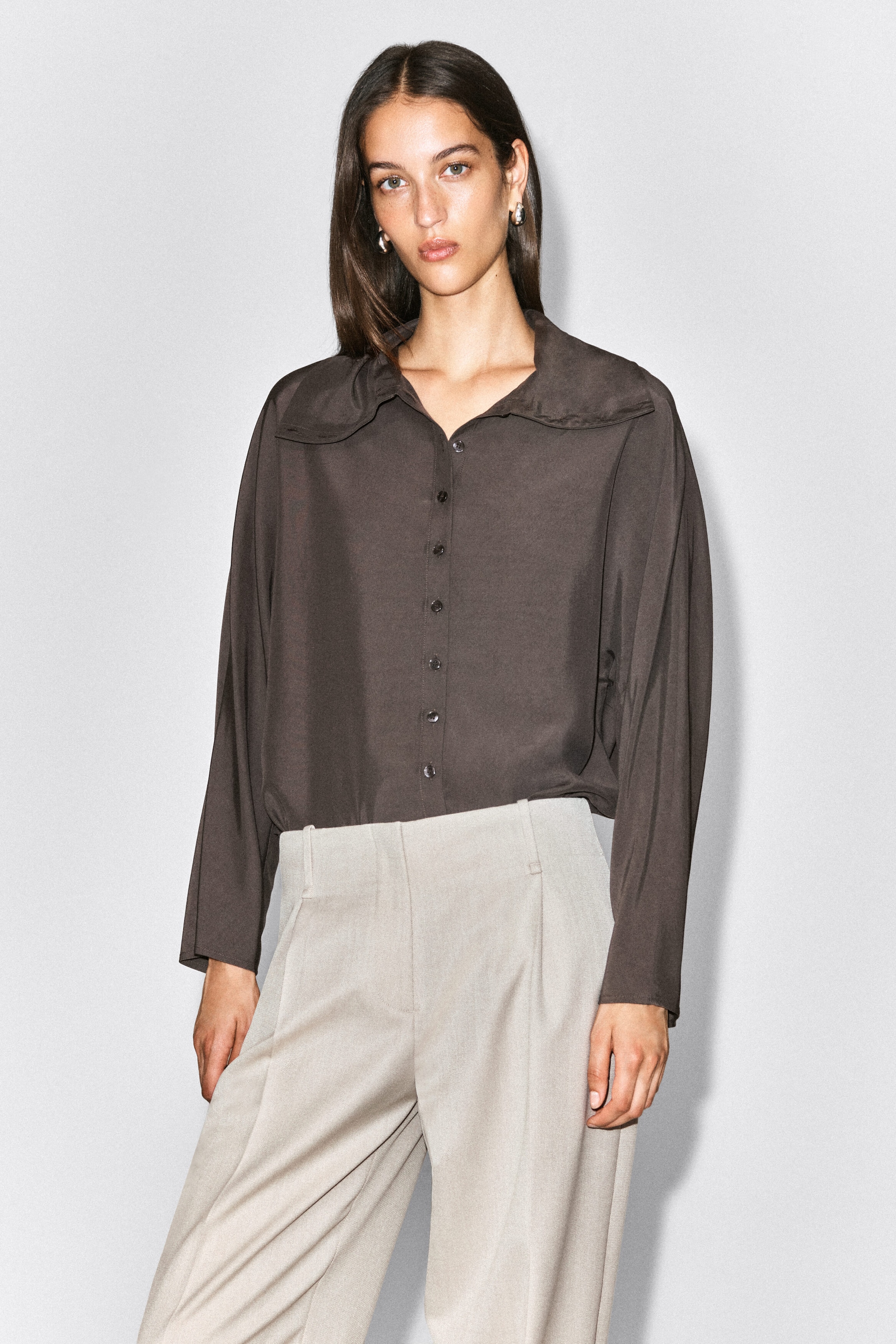 The model is wearing light beige pleated trousers featuring a clean waistband with belt loops and structured front pleats. A dark brown, long-sleeve button-down shirt is tucked into the waistband to highlight the trousers’ high-rise fit. The model is wearing light beige pleated trousers featuring a clean waistband with belt loops and structured front pleats. A dark brown, long-sleeve button-down shirt is tucked into the waistband to highlight the trousers’ high-rise fit.