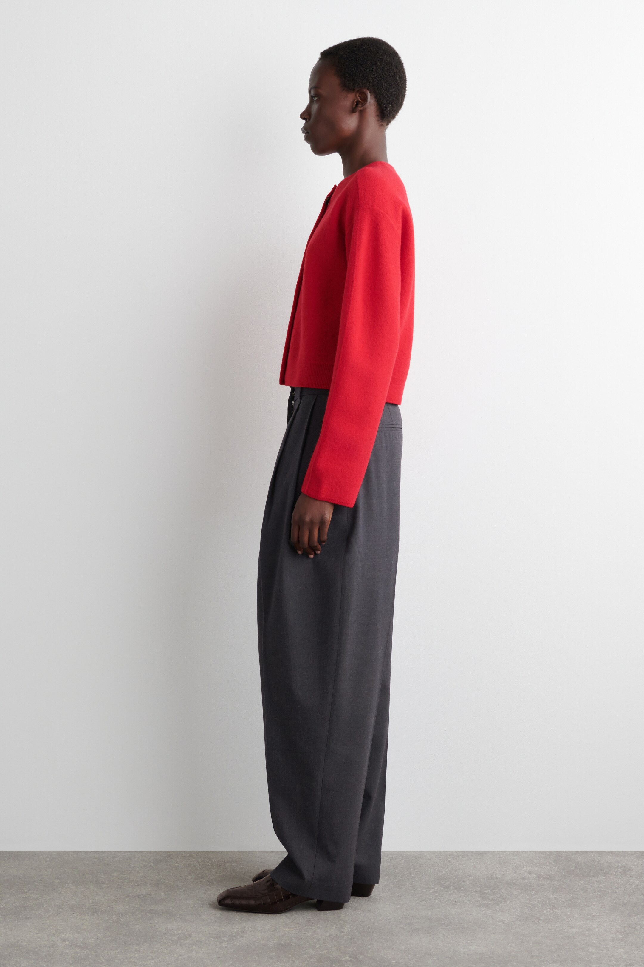 Agrandir l'image: Une veste courte, rouge, à manches longues est portée avec un pantalon gris à large jambe et plissé, l'ensemble étant présenté en profil latéral. La veste présente un décolleté rond et une silhouette carrée, tandis que le pantalon tombe au sol et couvre partiellement des chaussures marron foncé à bout pointu.