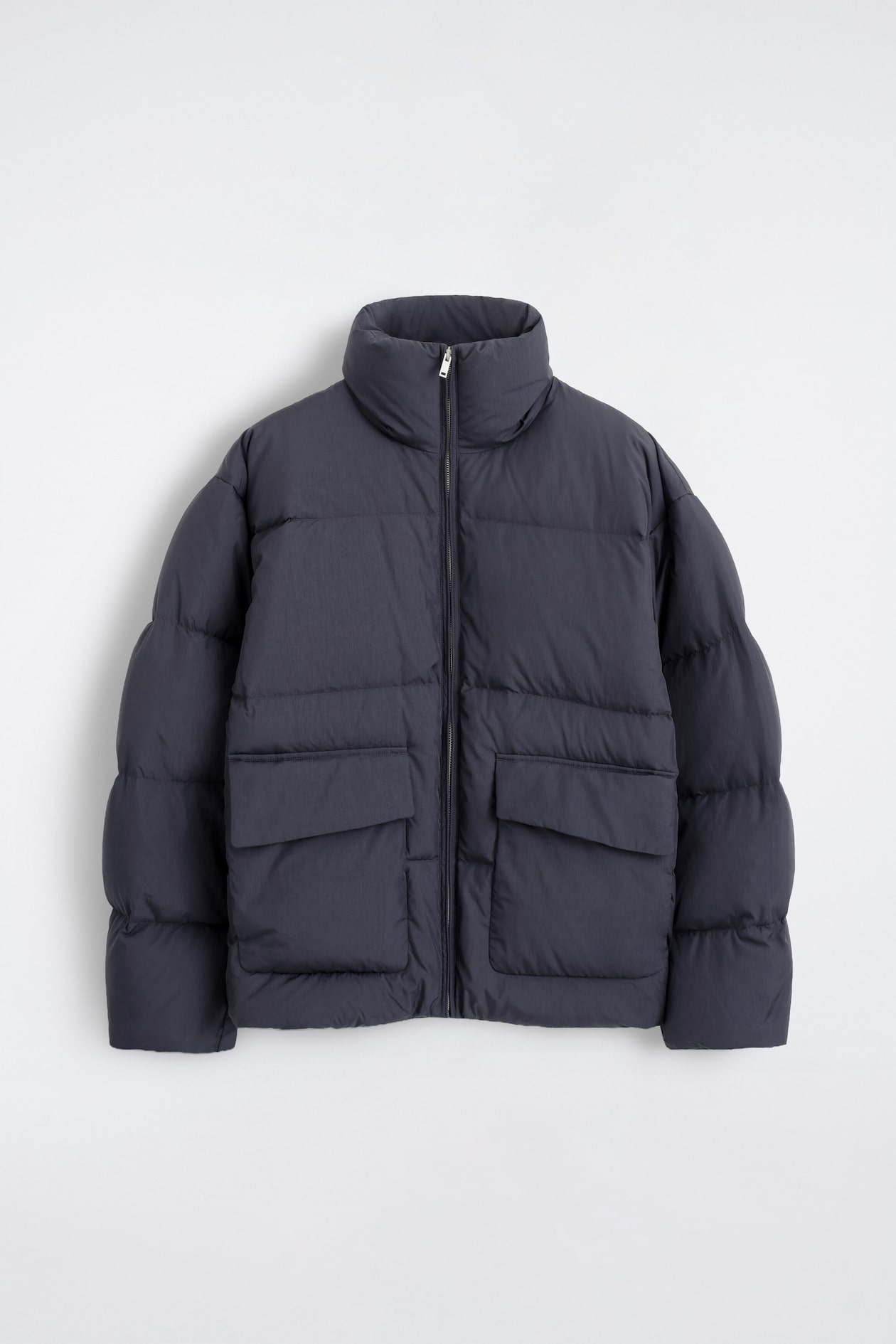 The funnel-neck nylon down puffer jacket is shown from the front, featuring a dark gray quilted design with a high funnel collar and a center zip closure. It features two large front flap pockets and puffy, segmented sleeves.