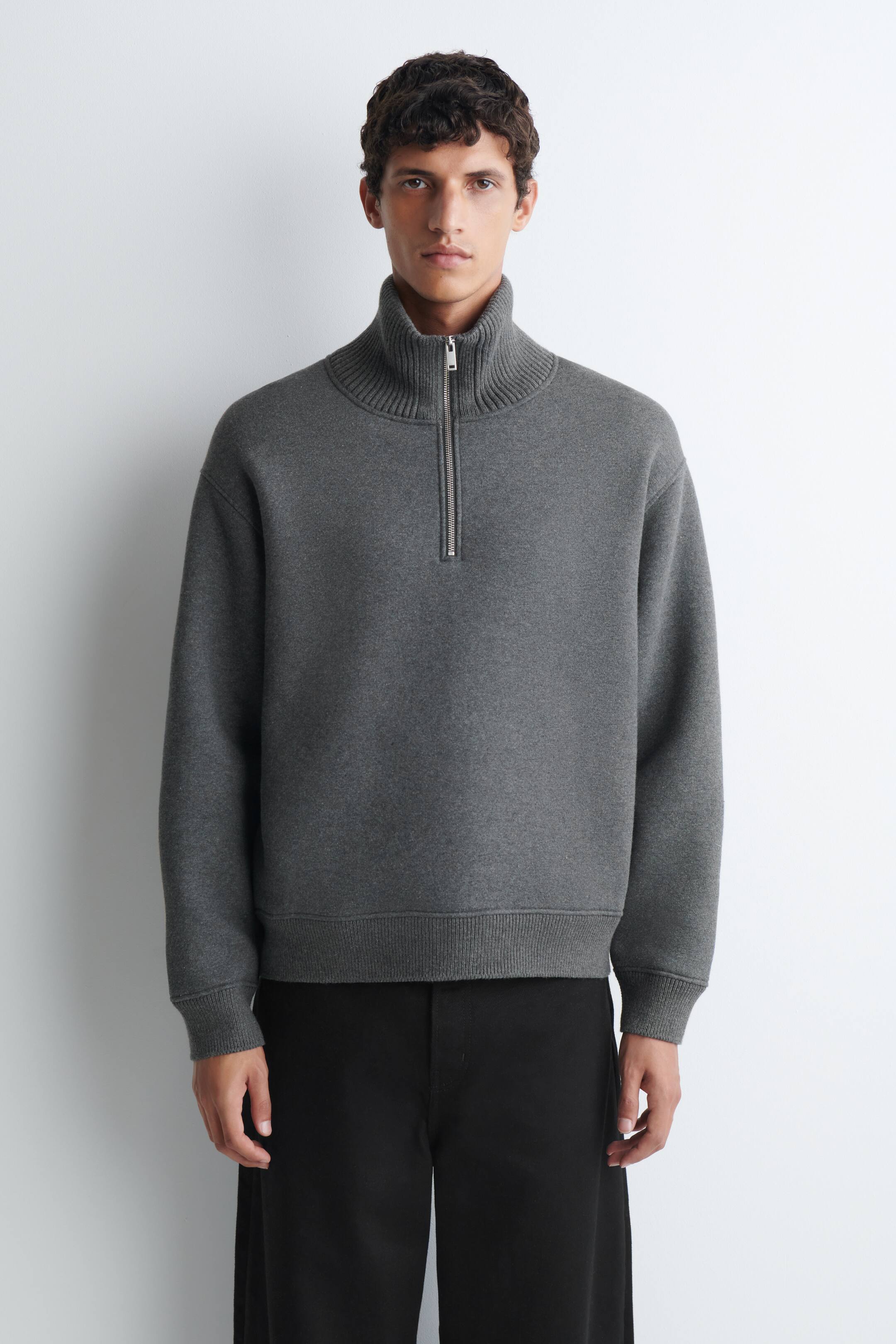 Agrandir l'image: Le mannequin porte un pull zippé en jersey de coton et de laine gris, doté d’un col montant côtelé avec une fermeture demi-zip de couleur argentée. Le pull est associé à un pantalon noir et présente des poignets côtelés ainsi qu’un ourlet côtelé.