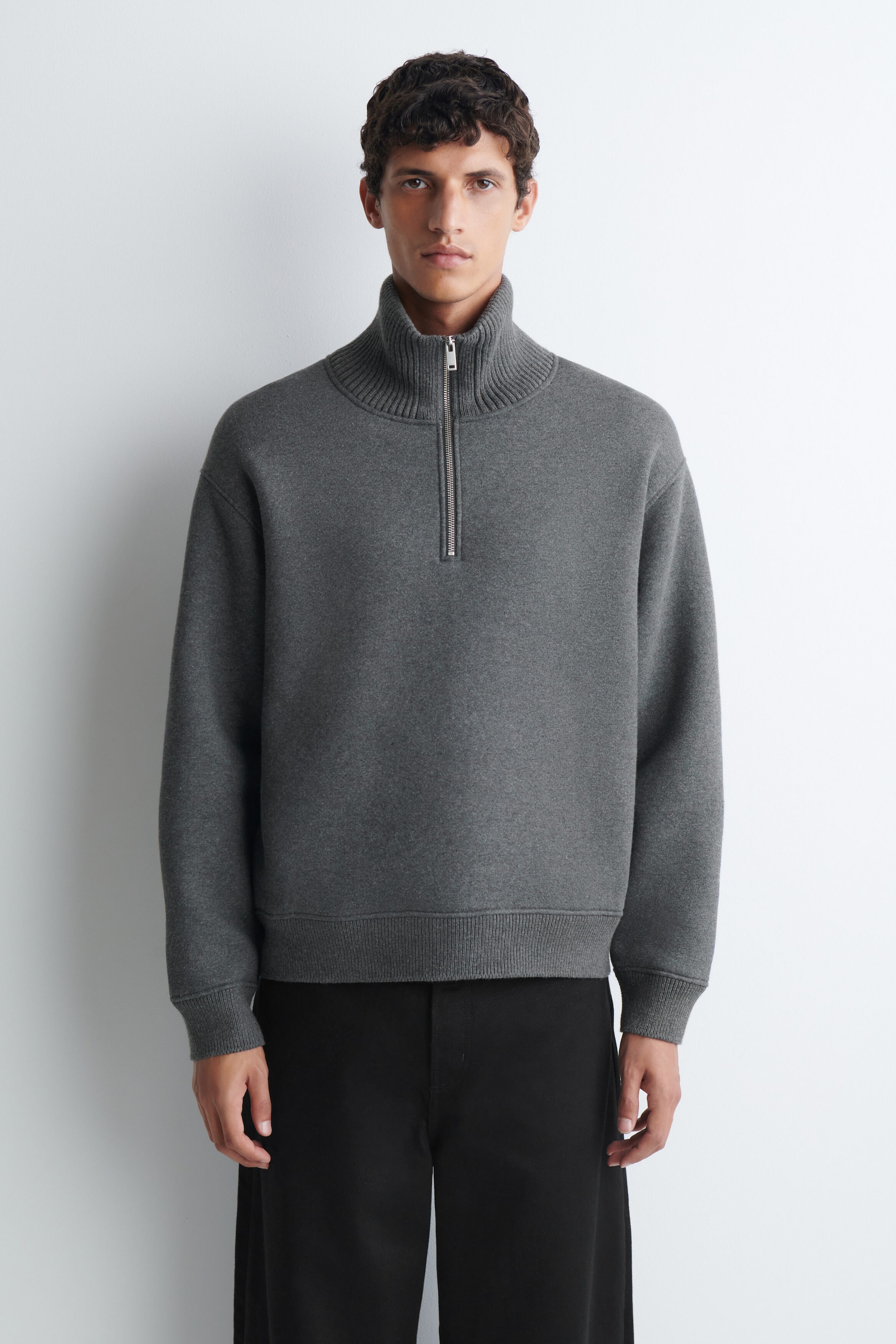 A grey cotton-blend jersey zip-up sweater is being worn by the model, featuring a high ribbed collar with a silver-tone half-zip closure. The sweater is styled with black trousers and has ribbed cuffs and a ribbed hem. A grey cotton-blend jersey zip-up sweater is being worn by the model, featuring a high ribbed collar with a silver-tone half-zip closure. The sweater is styled with black trousers and has ribbed cuffs and a ribbed hem.