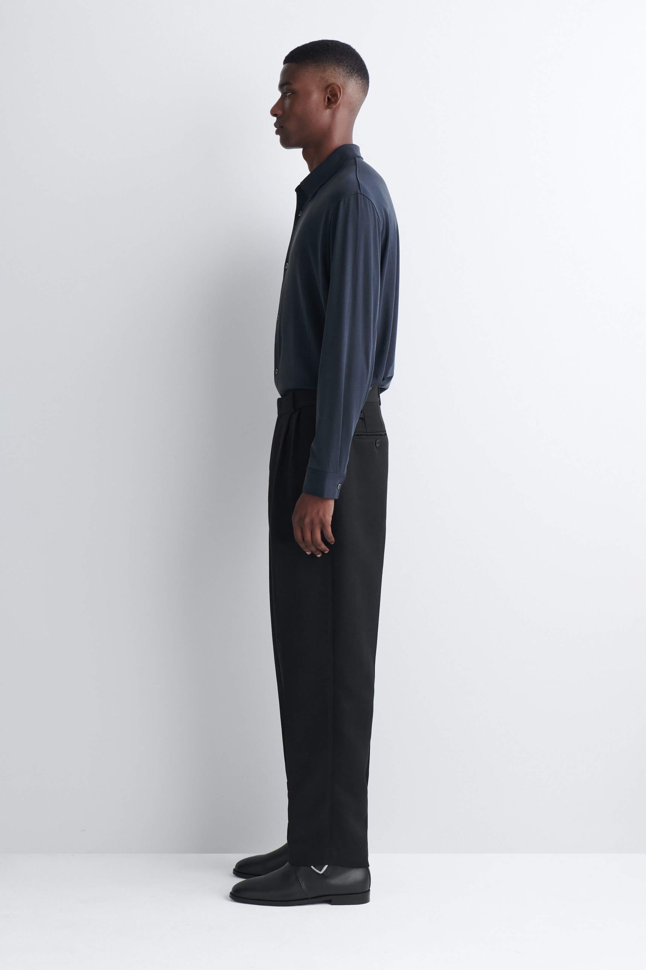 The model is wearing a dark gray fluid interlock jersey shirt in side profile, tucked into tailored black trousers. The long-sleeve shirt features a classic collar and buttoned cuffs, highlighting its soft drape and smooth texture. The model is wearing a dark gray fluid interlock jersey shirt in side profile, tucked into tailored black trousers. The long-sleeve shirt features a classic collar and buttoned cuffs, highlighting its soft drape and smooth texture.