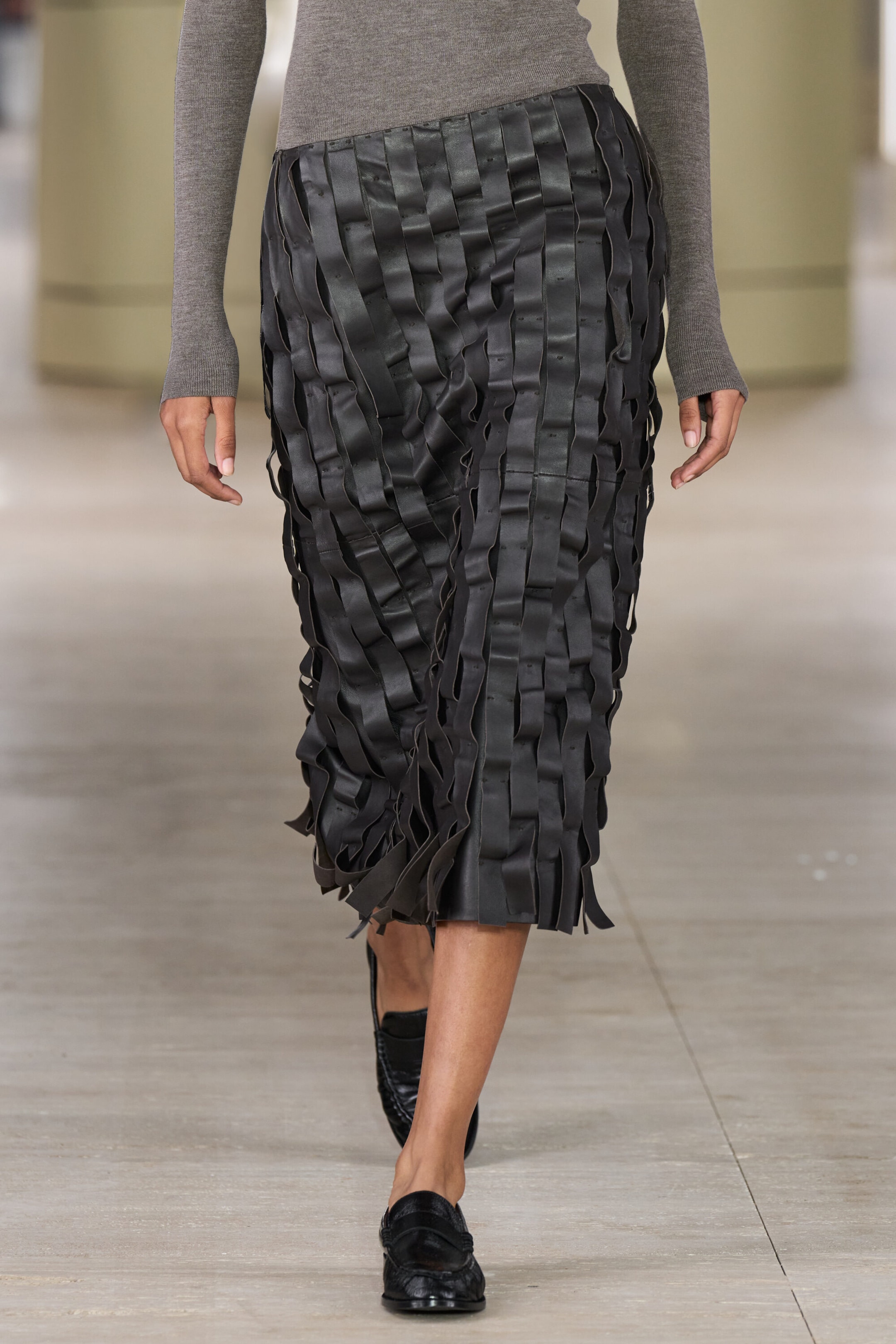 A raw-cut leather midi skirt is shown from the front, featuring vertical overlapping black leather strips that create a textured, woven design with loose, raw-cut ends at the hem. The slim-fit skirt sits at the waist and is styled with a gray long-sleeve knit top. A raw-cut leather midi skirt is shown from the front, featuring vertical overlapping black leather strips that create a textured, woven design with loose, raw-cut ends at the hem. The slim-fit skirt sits at the waist and is styled with a gray long-sleeve knit top.