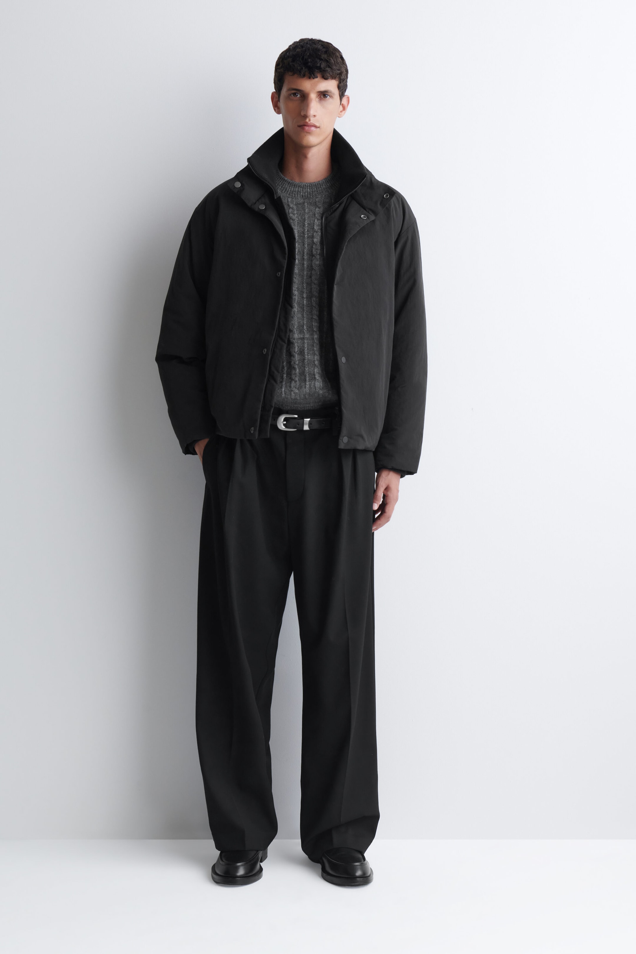 A black padded funnel-neck down jacket is being worn open by the model, showing off a high collar and snap-button closures. The jacket is layered over a textured grey knit sweater and black wide-leg pants. A black padded funnel-neck down jacket is being worn open by the model, showing off a high collar and snap-button closures. The jacket is layered over a textured grey knit sweater and black wide-leg pants.
