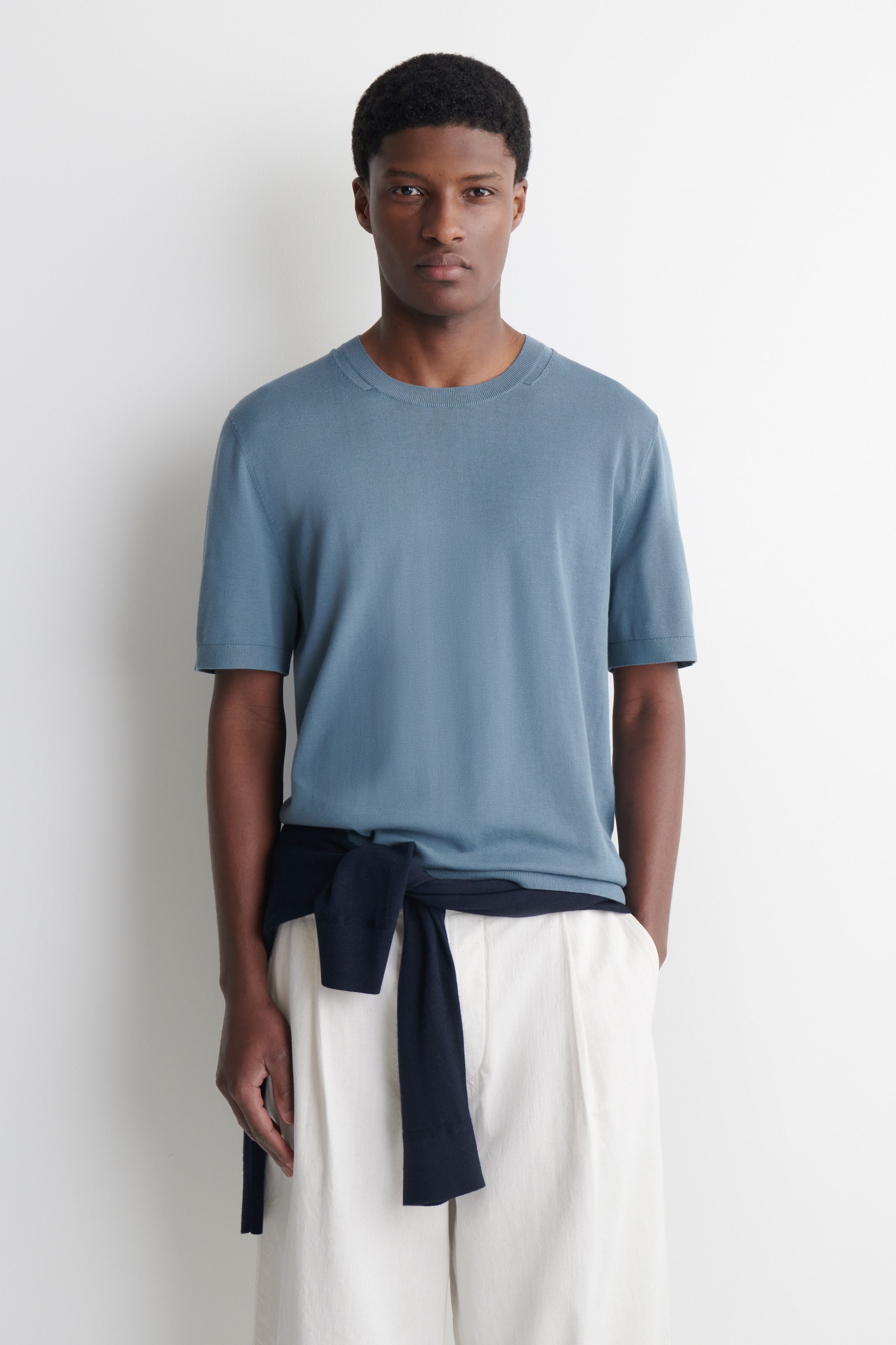 Größeres Bild ansehen: Das staubige blaue T-Shirt mit kurzen Ärmeln verfügt über einen gerippten Rundhalsausschnitt, eine glatte Feinstricktextur und eine entspannte Passform, die nach vorne zeigt.