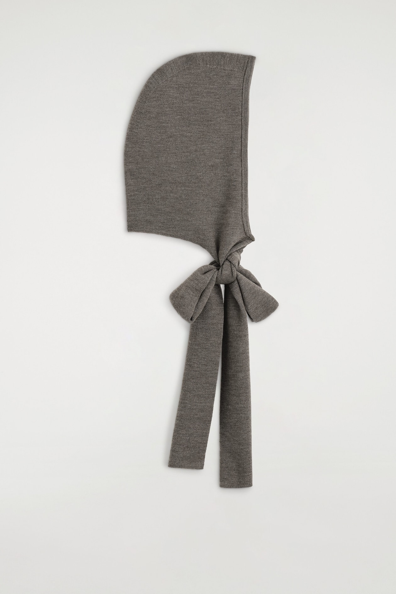 Ein fein gestrickter Schal mit Kapuze in Taupe-Grau wird in der seitlichen Profilansicht gezeigt. Das Design verfügt über eine strukturierte Kapuze mit langen, flachen Bändern, die am Ansatz zu einer Schleife gebunden sind und senkrecht herabhängen.