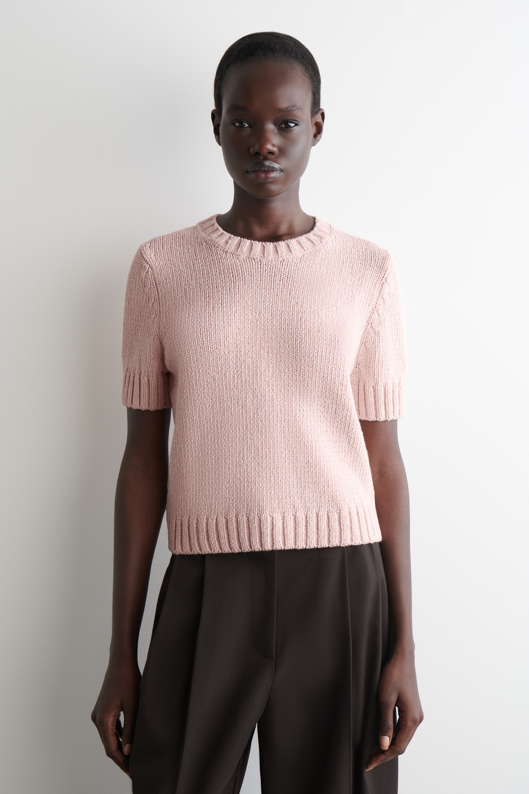 Größeres Bild anzeigen: Ein hellrosa kurzärmeliger Pullover wird präsentiert, der direkt nach vorne zeigt. Er verfügt über einen Rundhalsausschnitt, gerippte Ärmelbündchen und einen gerippten geraden Saum und ist in regulärer Passform geschnitten, an der Taille abgeschnitten.