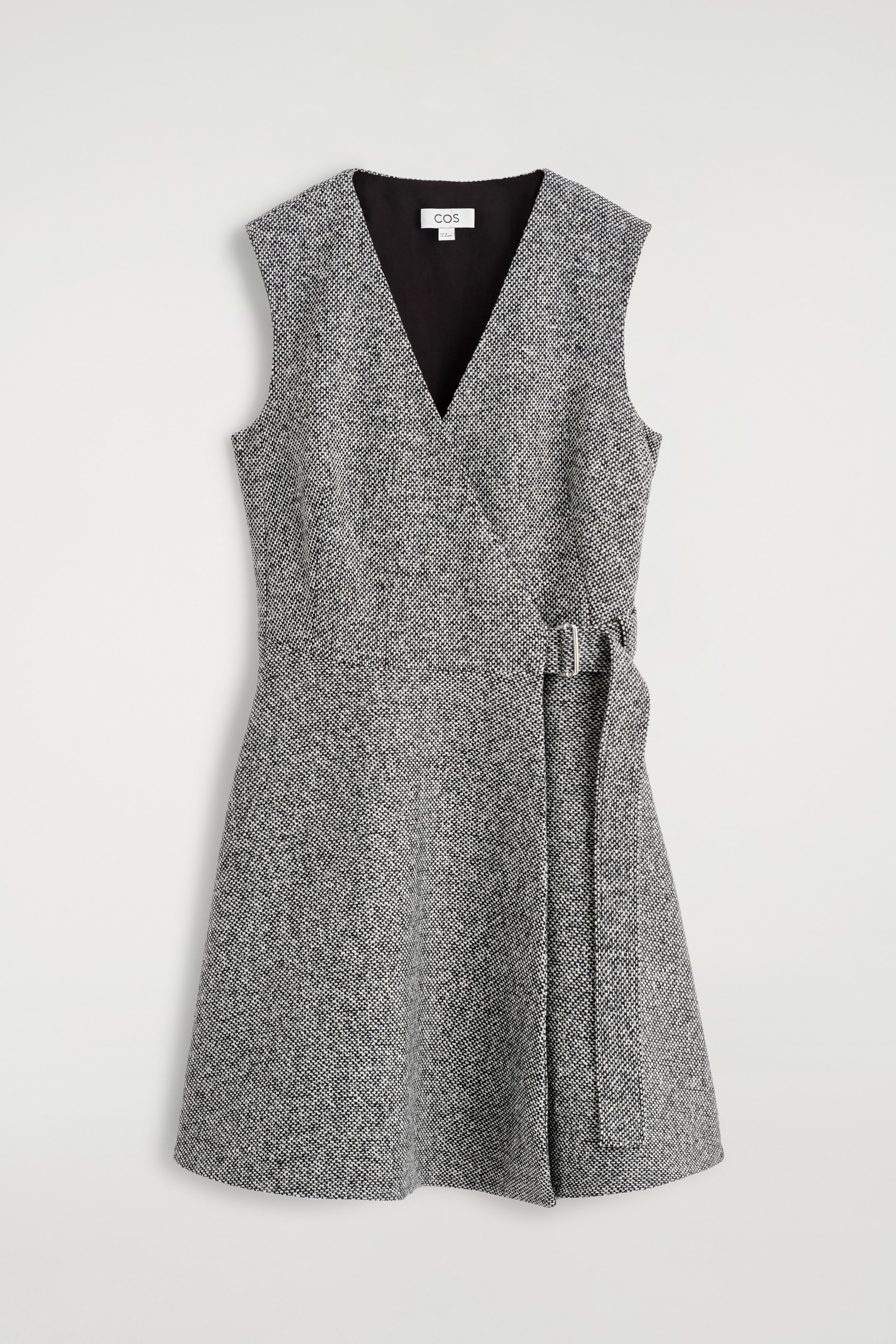 Ingrandisci l'immagine: Un mini abito in tweed ricco di texture in bianco e nero è presentato frontalmente, con un scollo a V e una forma strutturata a trapezio. Una cintura con fibbia stringe la vita sul lato sinistro della portatrice, e una fodera nera è visibile allo scollo a V.