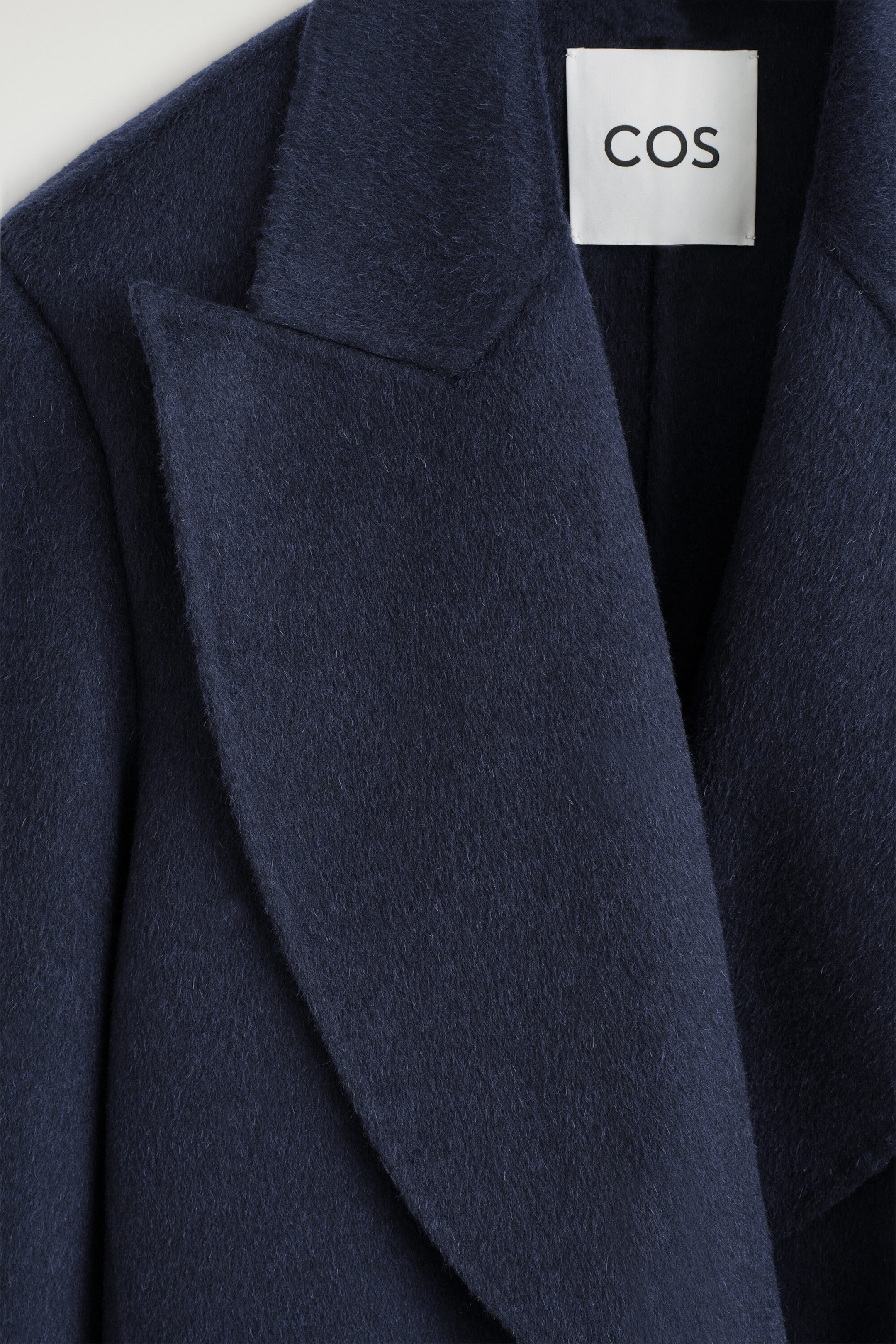 A navy blue wool-blend blazer is shown in a close-up front view, highlighting its soft, brushed texture and wide draped lapel. A white square brand label with the text 'COS' is visible on the inner back collar.