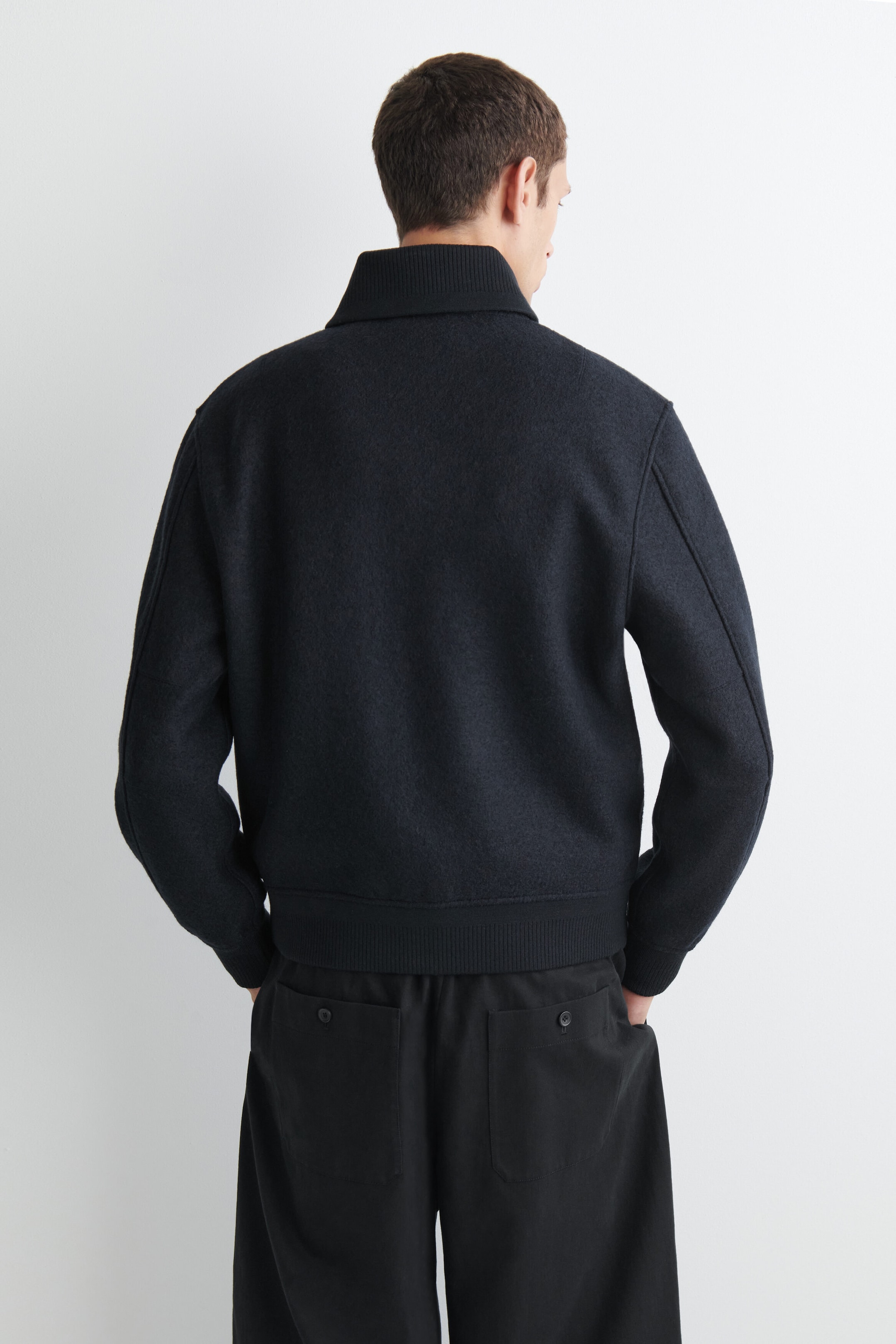 The boiled-wool shawl-collar jacket is shown from the back, highlighting its textured dark navy fabric and structured fit. The design features a high ribbed collar, ribbed cuffs, and a matching ribbed hem that rests at the waist of black trousers. The boiled-wool shawl-collar jacket is shown from the back, highlighting its textured dark navy fabric and structured fit. The design features a high ribbed collar, ribbed cuffs, and a matching ribbed hem that rests at the waist of black trousers.