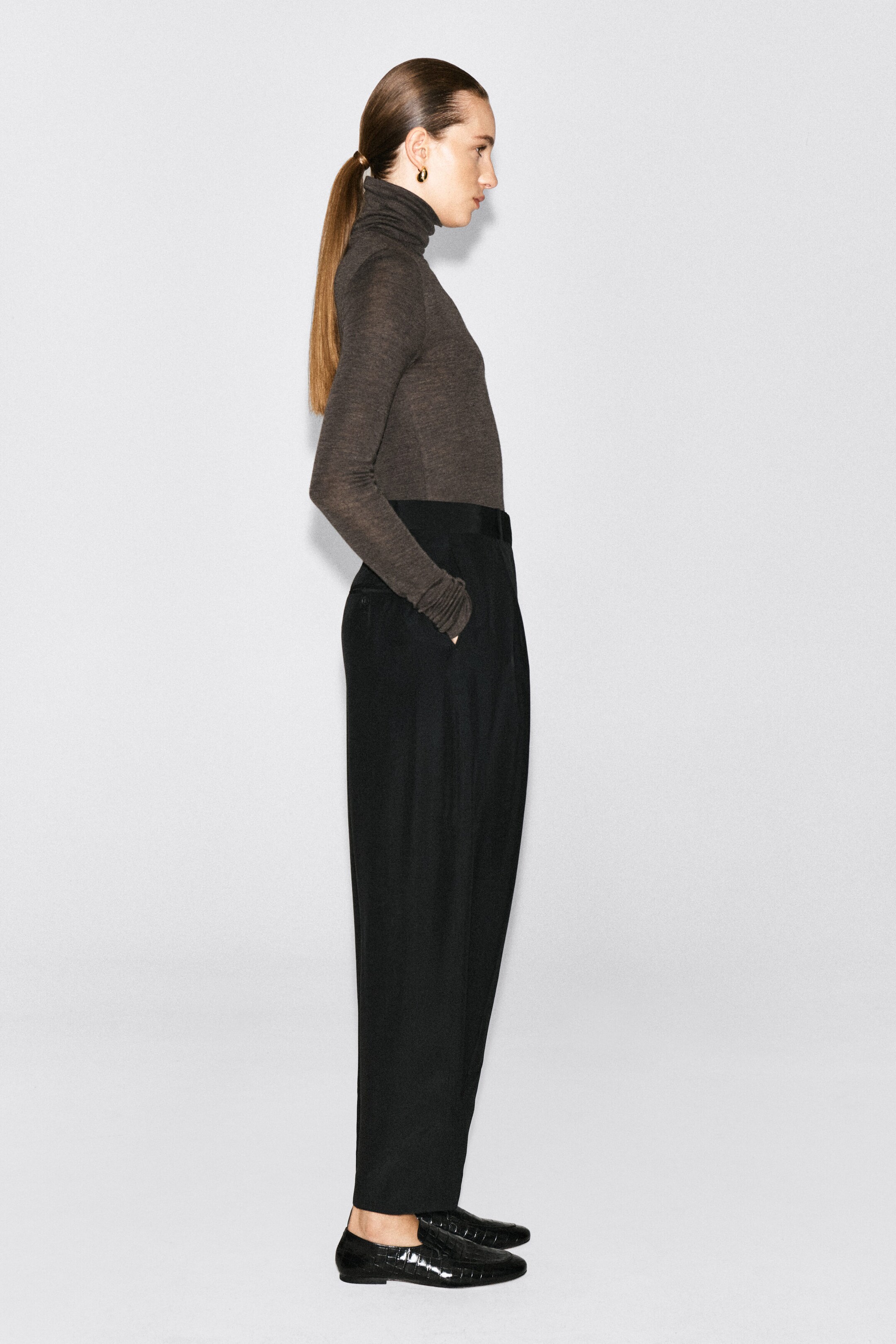 A merino wool turtleneck top in dark grey is being worn by the model, who is standing in profile. The long-sleeved, fine-knit top is tucked into high-waisted black trousers. A merino wool turtleneck top in dark grey is being worn by the model, who is standing in profile. The long-sleeved, fine-knit top is tucked into high-waisted black trousers.