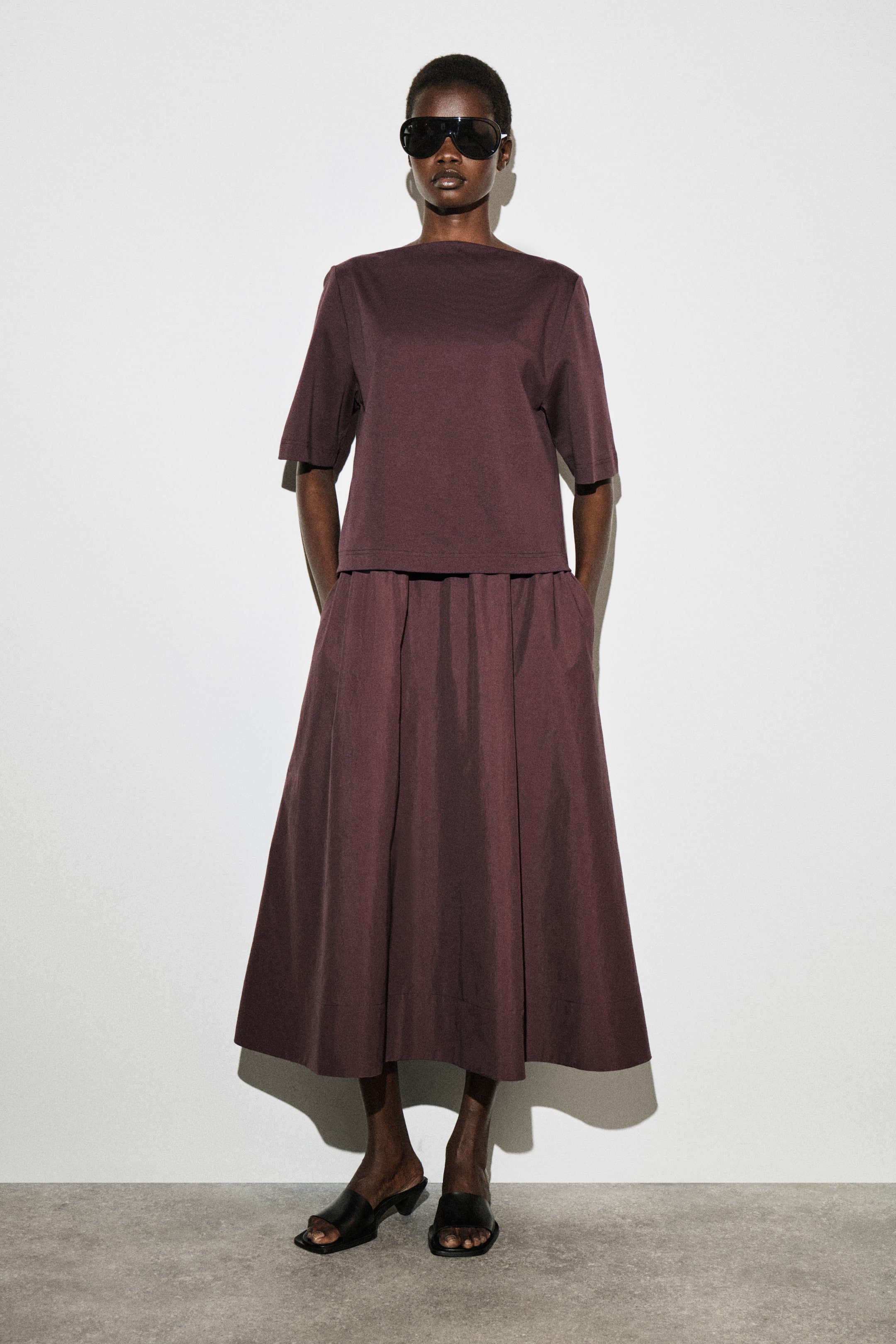 Größeres Bild anzeigen: Ein dunkelpflaumenfarbenes Zweiteiler-Set wird von vorne gezeigt, bestehend aus einem kurzärmeligen Oberteil mit Bootsausschnitt und einem passenden Midirock. Das Oberteil fällt locker, und der Rock sammelt sich an der Taille mit sichtbaren Seitentaschen, wodurch eine volle Silhouette entsteht.