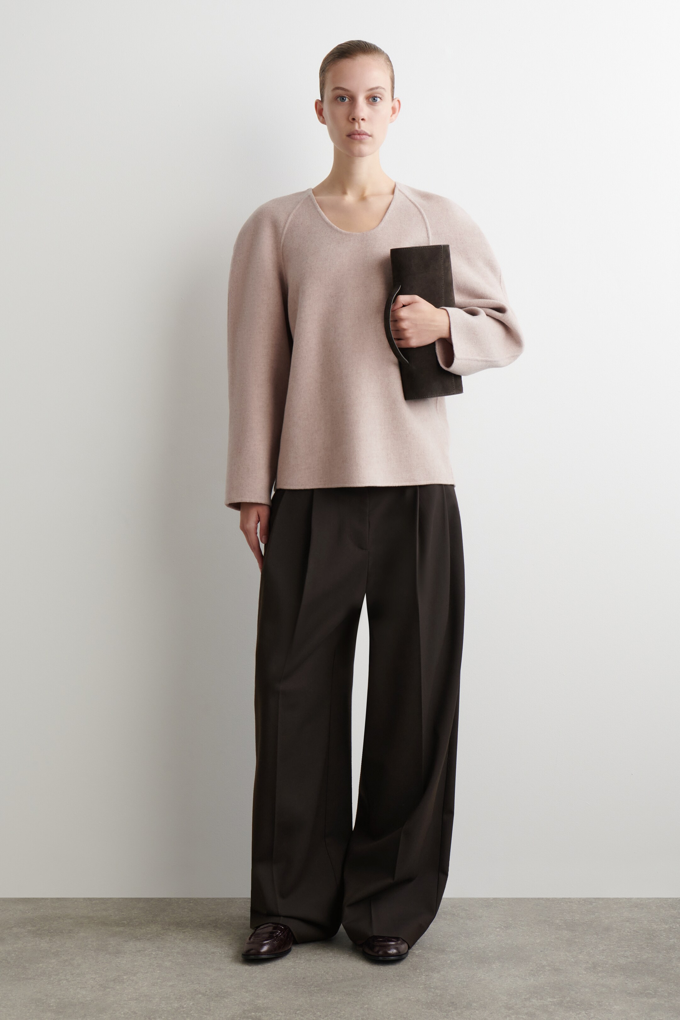 Grotere afbeelding bekijken: Een outfit wordt gepresenteerd, met een lichtroze, loszittende top met een ronde halslijn en raglan naden, gecombineerd met donkerbruine wijde plooibroek. Een donkerbruine clutch met een handvat wordt in de linkerhand gehouden, en er worden donkerbruine loafers gedragen.