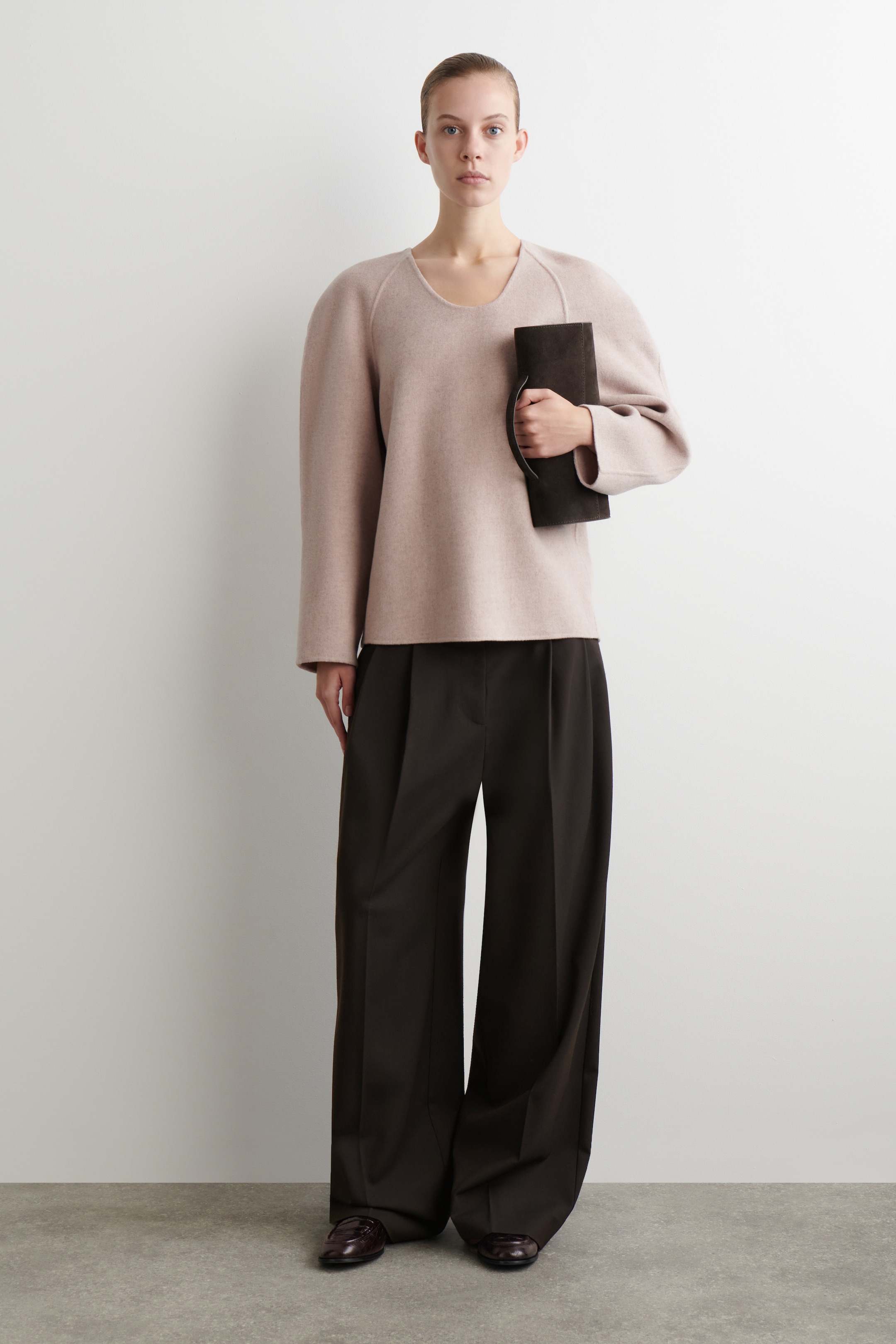 View larger image: An outfit is being presented, featuring a pale pink, loose-fitting top with a scoop neckline and raglan seams, paired with dark brown wide-leg pleated trousers. A dark brown clutch bag with a handle is being held in the left hand, and dark brown loafers are being worn.