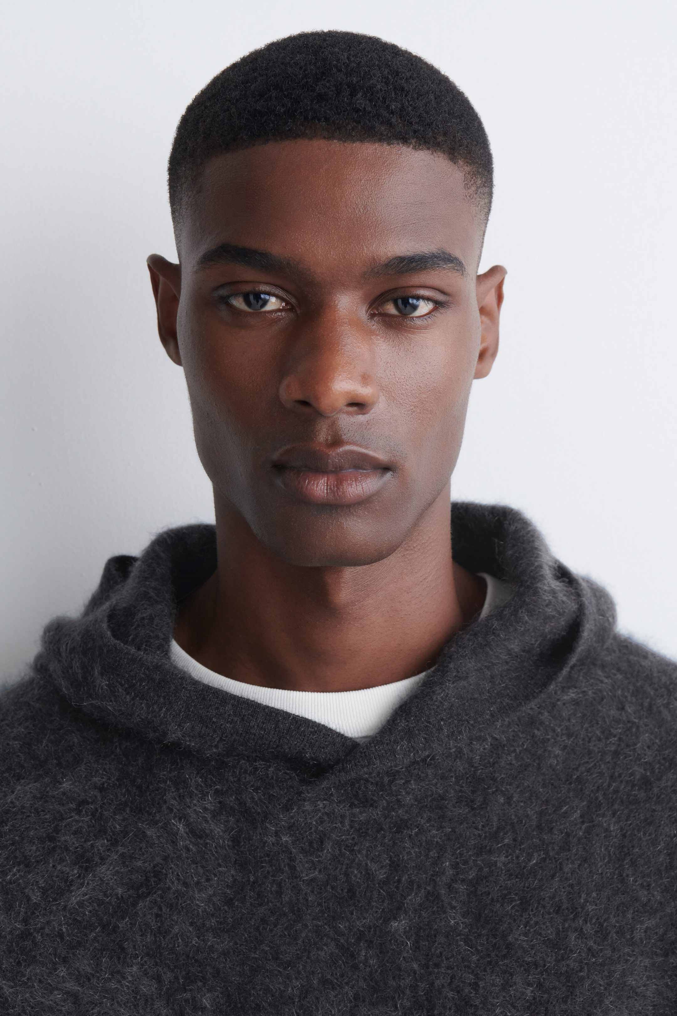 A dark gray brushed cashmere hoodie with a soft, textured finish and a relaxed hood is being worn by the model. The hoodie is layered over a visible white crew-neck T-shirt.