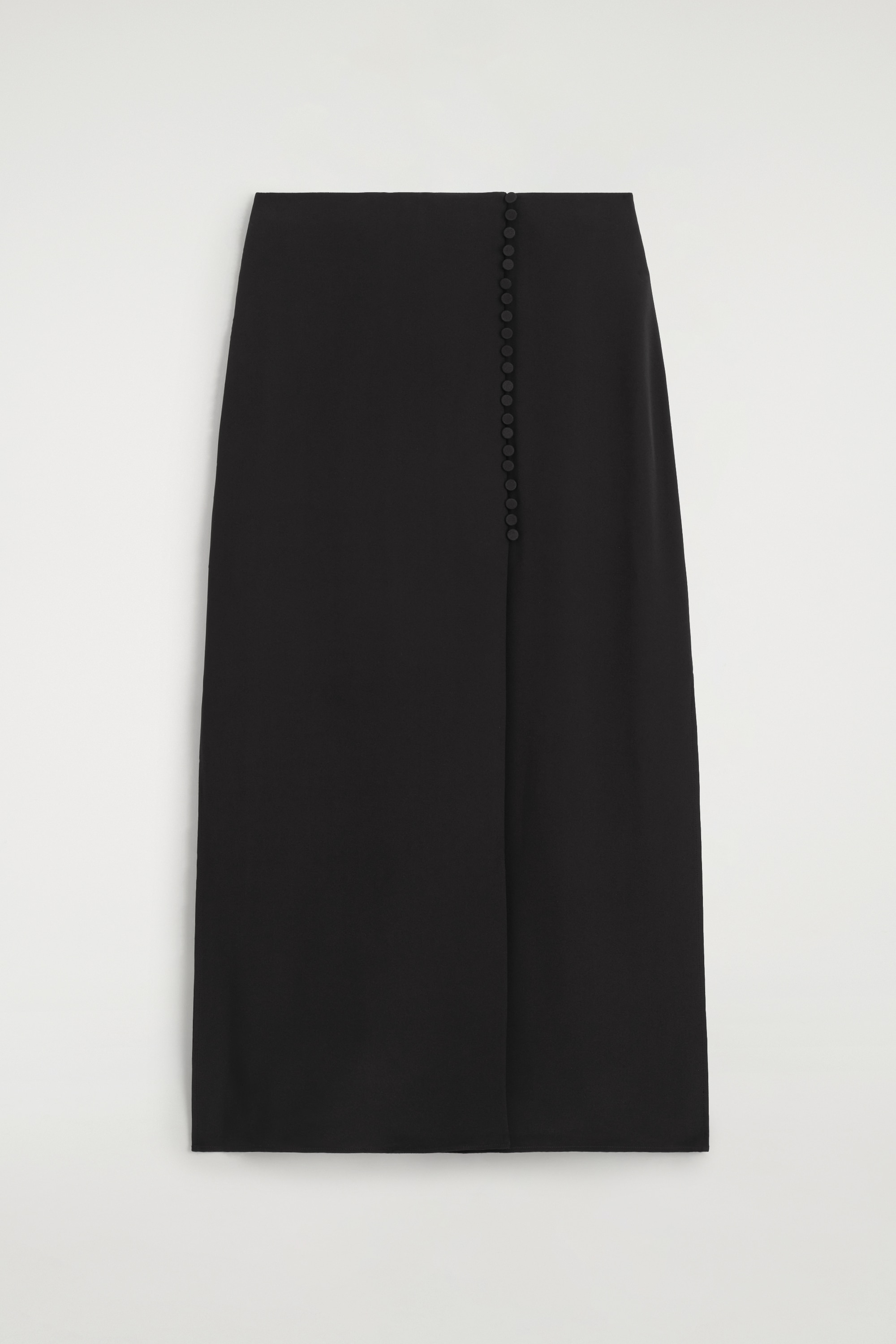 Le modÃ¨le porte un chemisier noir Ã  col montant et manches longues, avec des boutons sur le devant, ainsi quâune jupe longue assortie. Le modÃ¨le tient dâune main un petit sac Ã  main noir en cuir et porte des chaussures noires Ã  bout pointu.