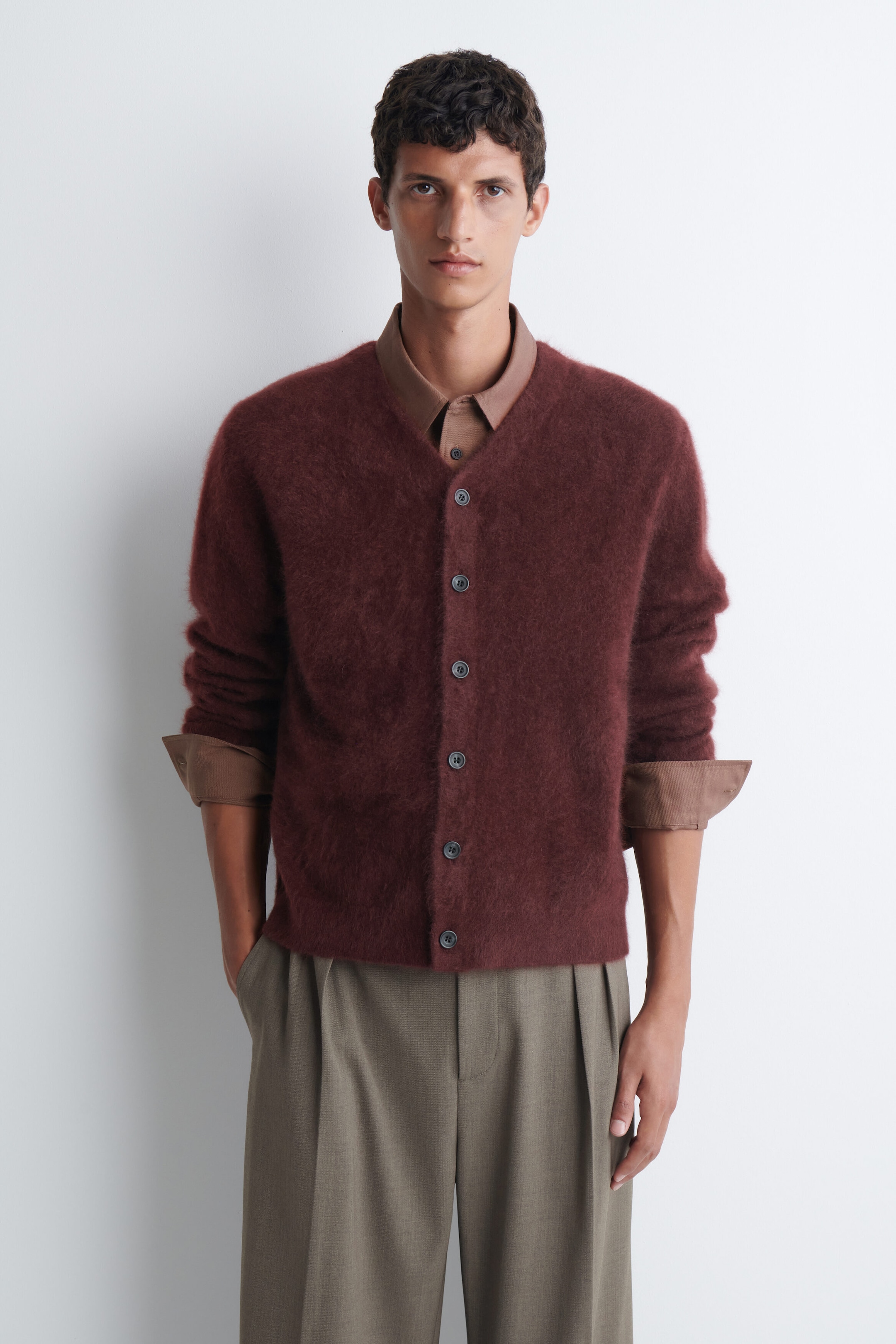 A burgundy brushed-cashmere V-neck cardigan featuring a button-front fastening is worn by the model. The cardigan is layered over a brown collared shirt with turned-up cuffs and styled with pleated olive-green trousers. A burgundy brushed-cashmere V-neck cardigan featuring a button-front fastening is worn by the model. The cardigan is layered over a brown collared shirt with turned-up cuffs and styled with pleated olive-green trousers.