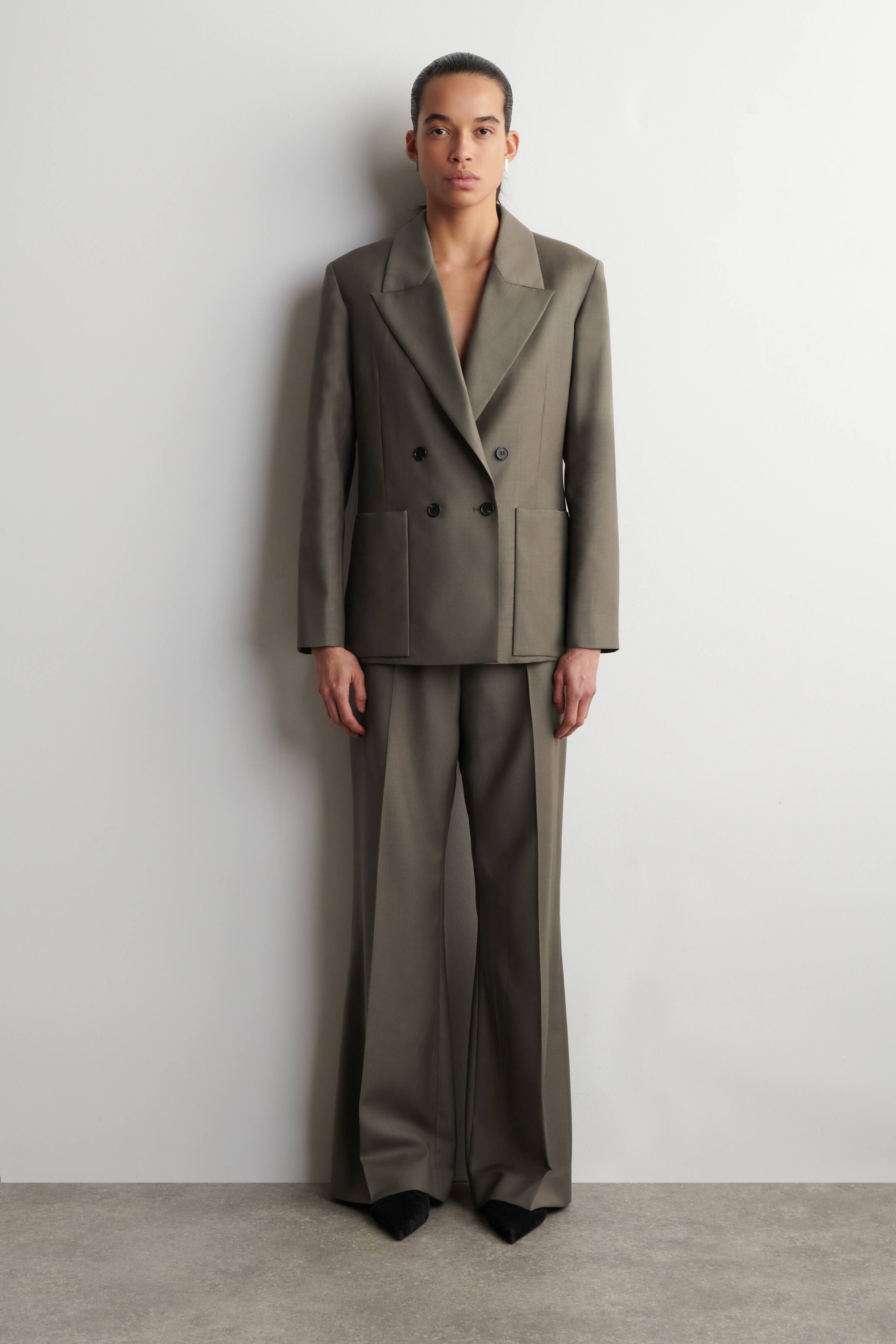 Agrandir l'image: Un tailleur-pantalon vert kaki est présenté, face avant. Le blazer croisé présente des épaules structurées, des revers crantés, quatre boutons sombres et deux grandes poches plaquées, tandis que le pantalon assorti à jambes larges affiche des plis marqués à l'avant.