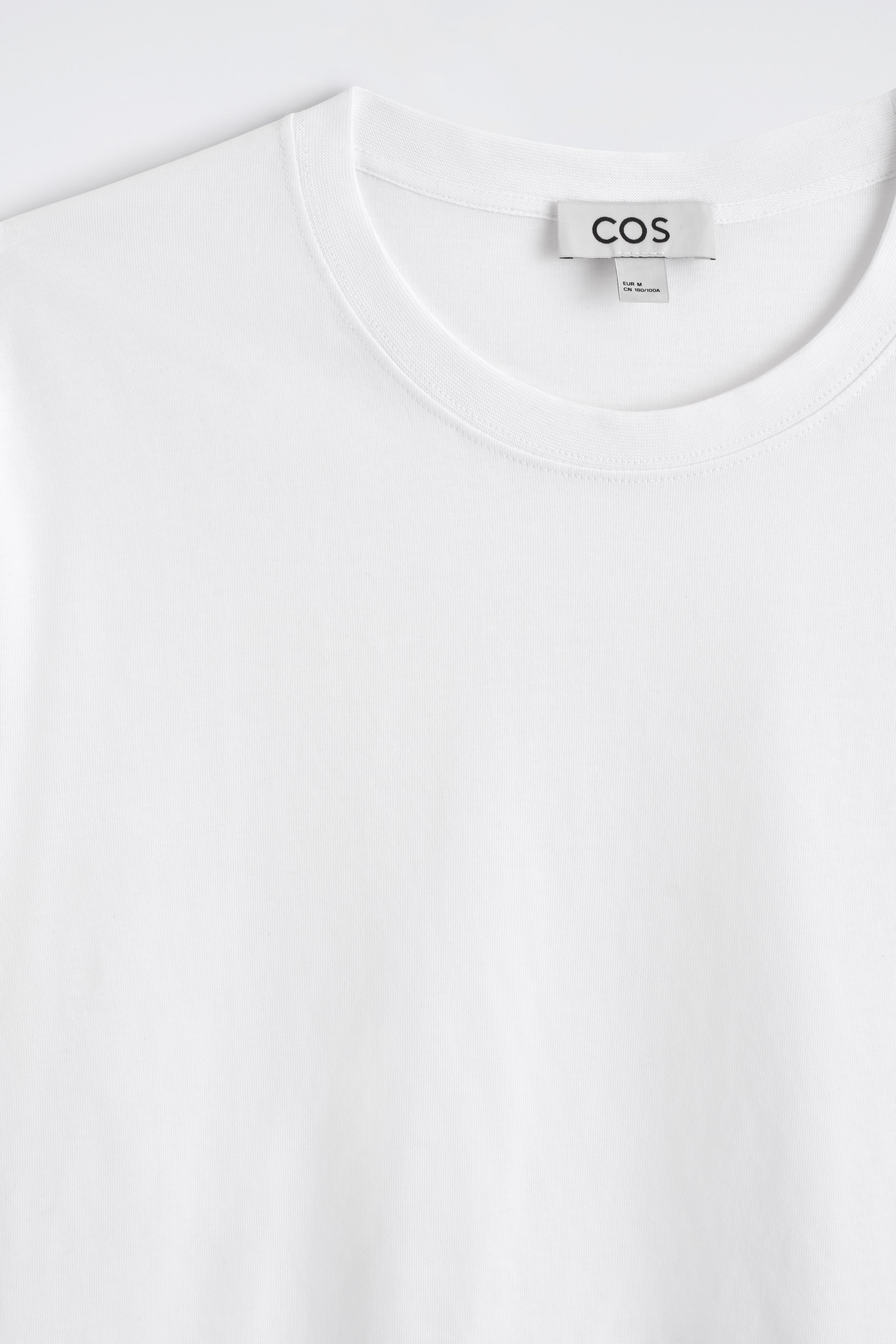 Grotere afbeelding bekijken: Een witte t-shirt ligt plat, met een vooraanzicht dat een ronde crew neck en een geribde kraag toont. De gladde stof vertoont een fijne breistructuur, en een zwart-wit merklabel met de tekst 'COS' is zichtbaar aan de achterkant van de nek.