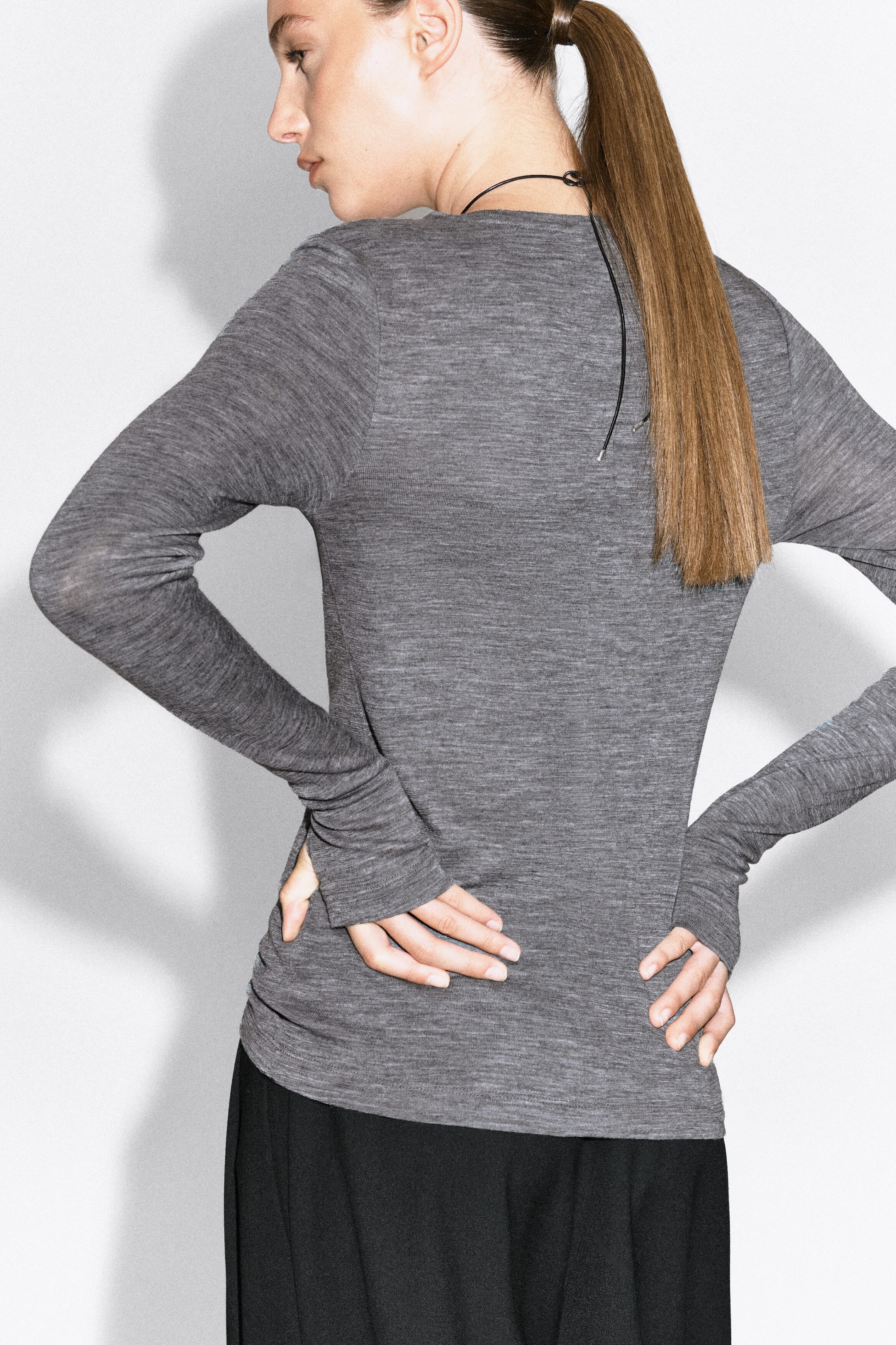 A slim merino wool top in textured grey is being worn by the model, who is standing with hands resting on the lower back to showcase the fitted silhouette and long sleeves. The long-sleeved top is paired with black trousers.