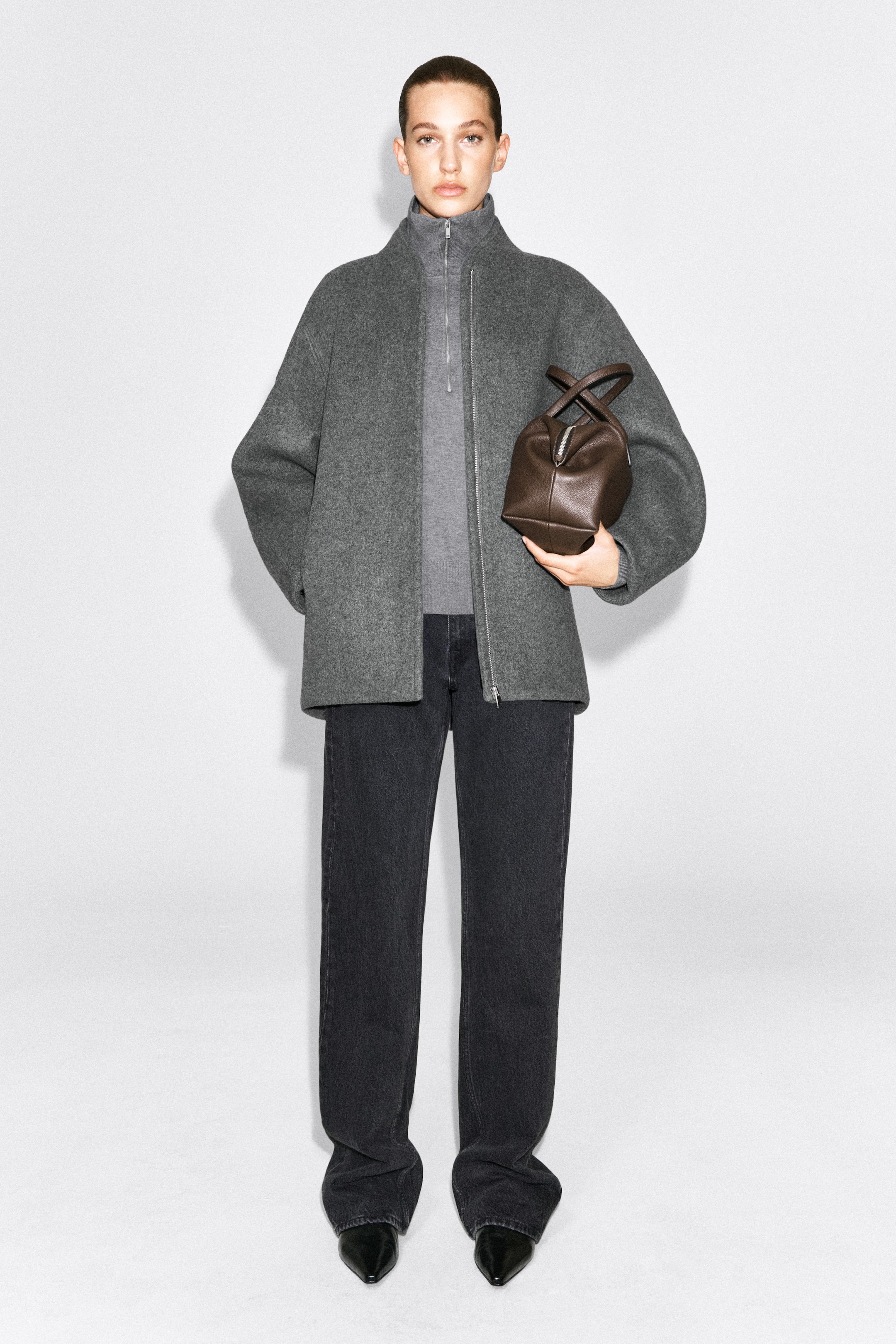 Visualizza immagine più grande: Una giacca con zip in misto lana scolpita, grigia, indossata dalla modella, caratterizzata da una silhouette oversize, collo alto e zip frontale argentata. La giacca è portata aperta sopra un top half-zip grigio più chiaro ed è abbinata a jeans grigio scuro a gamba larga.