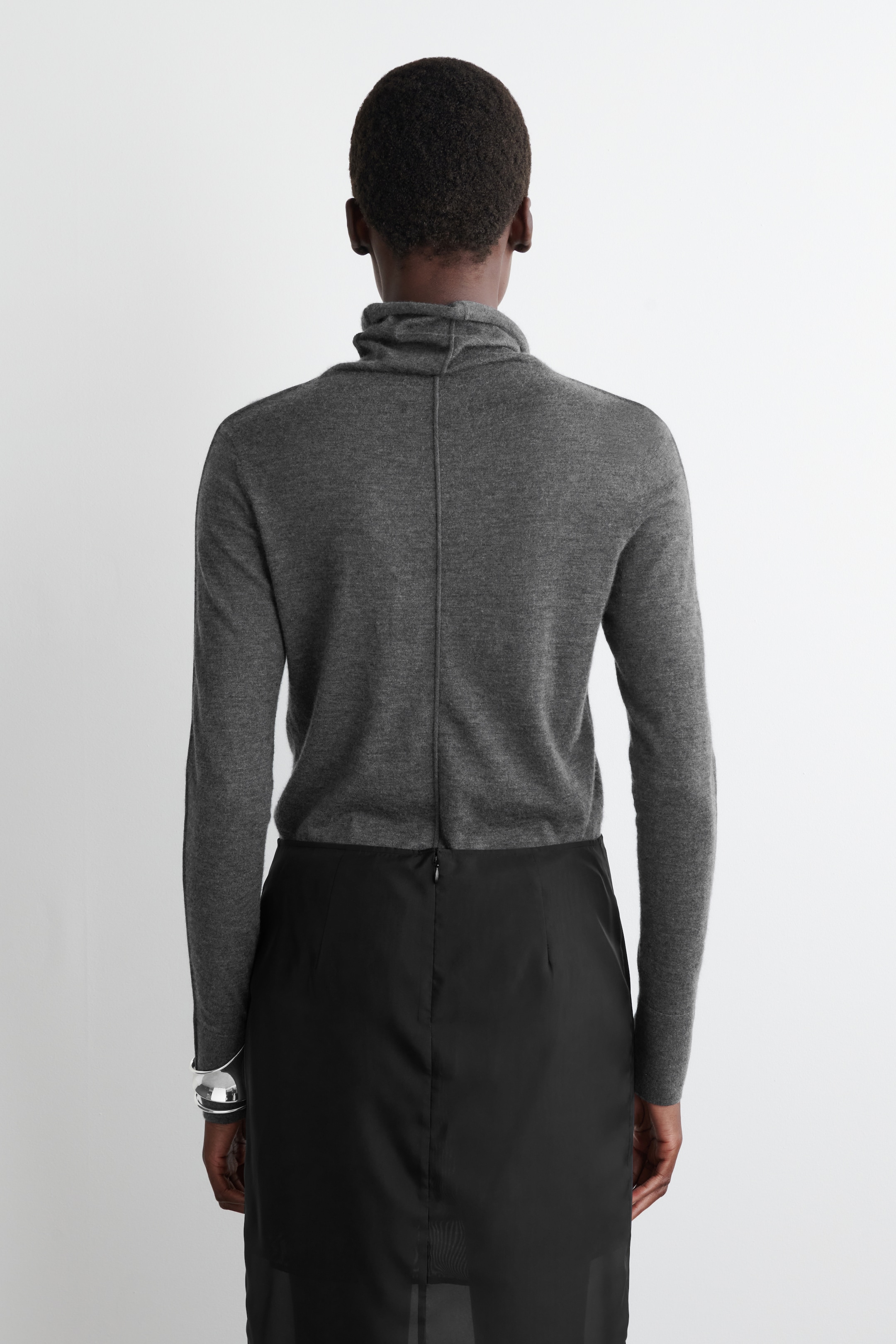 Grösseres Bild ansehen: Ein grauer drapierter Rollkragenpullover wird von hinten gezeigt, mit einer markanten vertikalen Naht, die in der Mitte verläuft. Er wird mit einem schwarzen Rock getragen, der einen sichtbaren Reißverschluss am Rücken hat, und ein silbernes Manschettenarmband schmückt das linke Handgelenk.