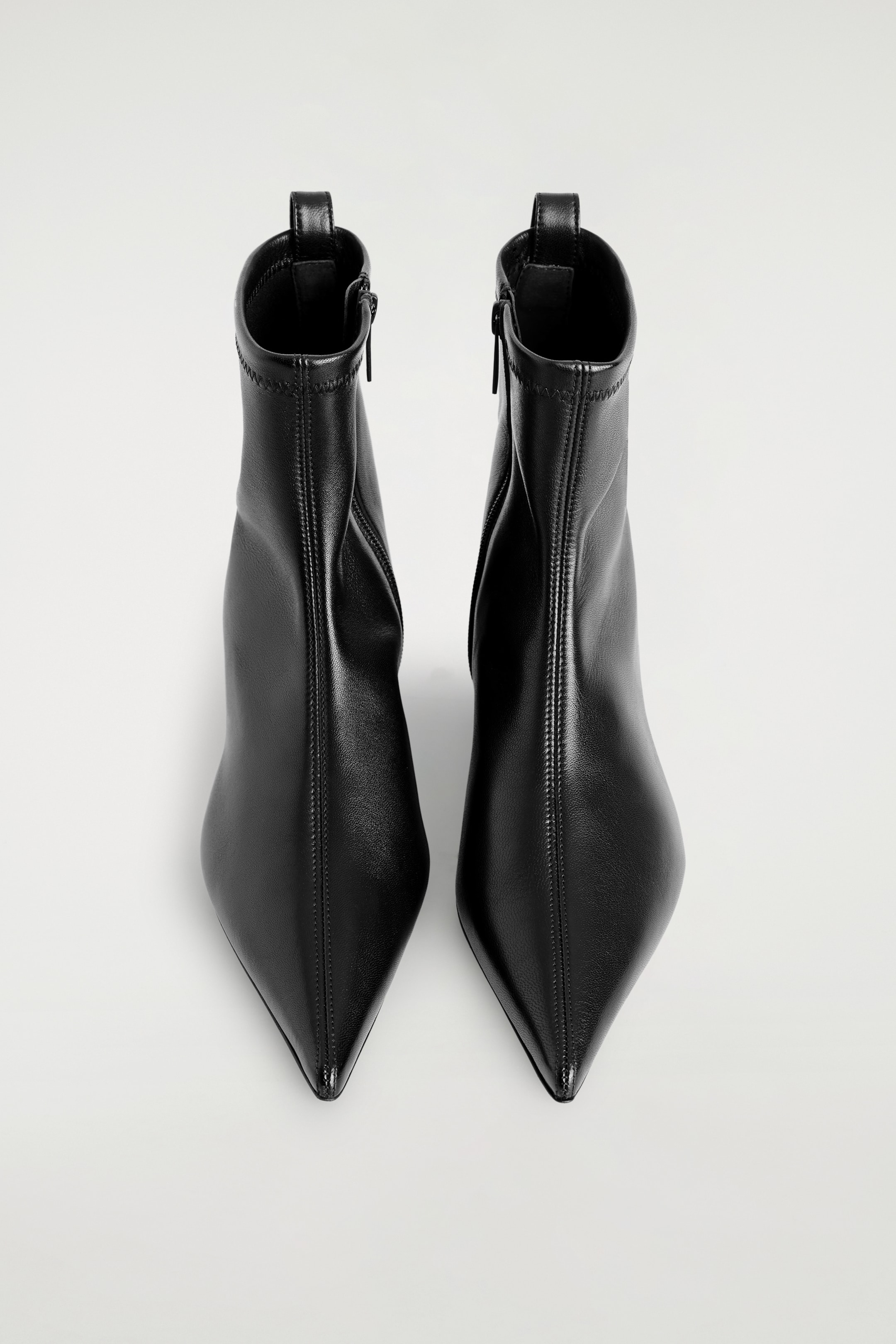 Agrandir l'image: Deux bottines en cuir noir à bout pointu sont debout, face à l'avant. Chaque botte présente un bout pointu acéré, une couture verticale centrale proéminente et une tige ajustée à hauteur de cheville. La tige est détaillée avec une fermeture éclair sur le côté intérieur et une petite languette de tirage sur le bord supérieur arrière.