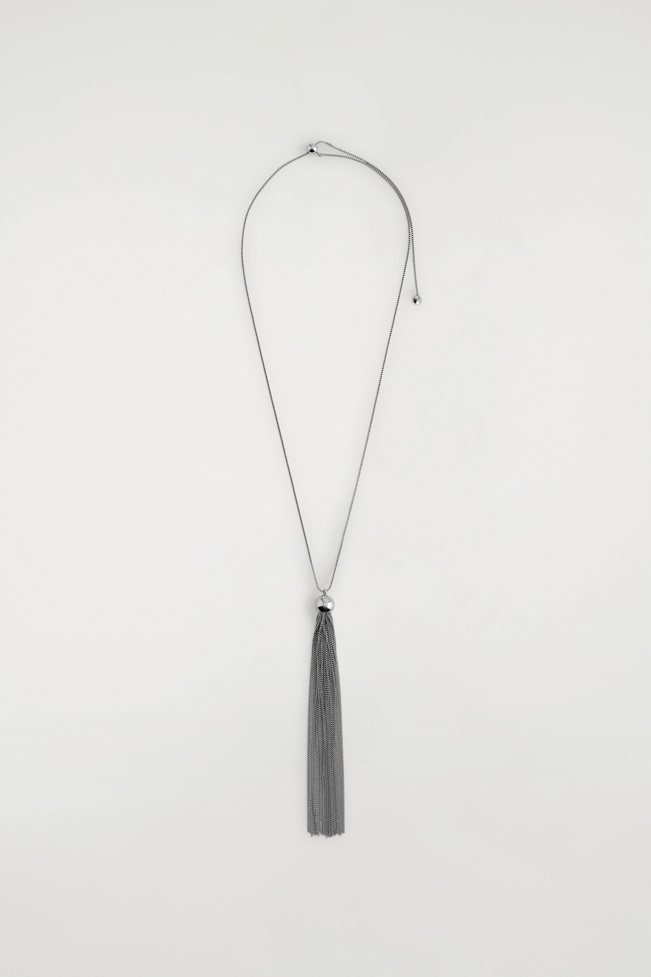 Ver imagen más grande: Un collar de tono metalizado está colocado plano, mostrando su parte frontal. Una cadena delgada con una cuenta deslizante ajustable y dos pequeños extremos con bolas forma el collar. Un colgante largo de borla está suspendido de la cadena; se compone de una pequeña cuenta esférica en su parte superior y numerosas cadenas finas colgantes.