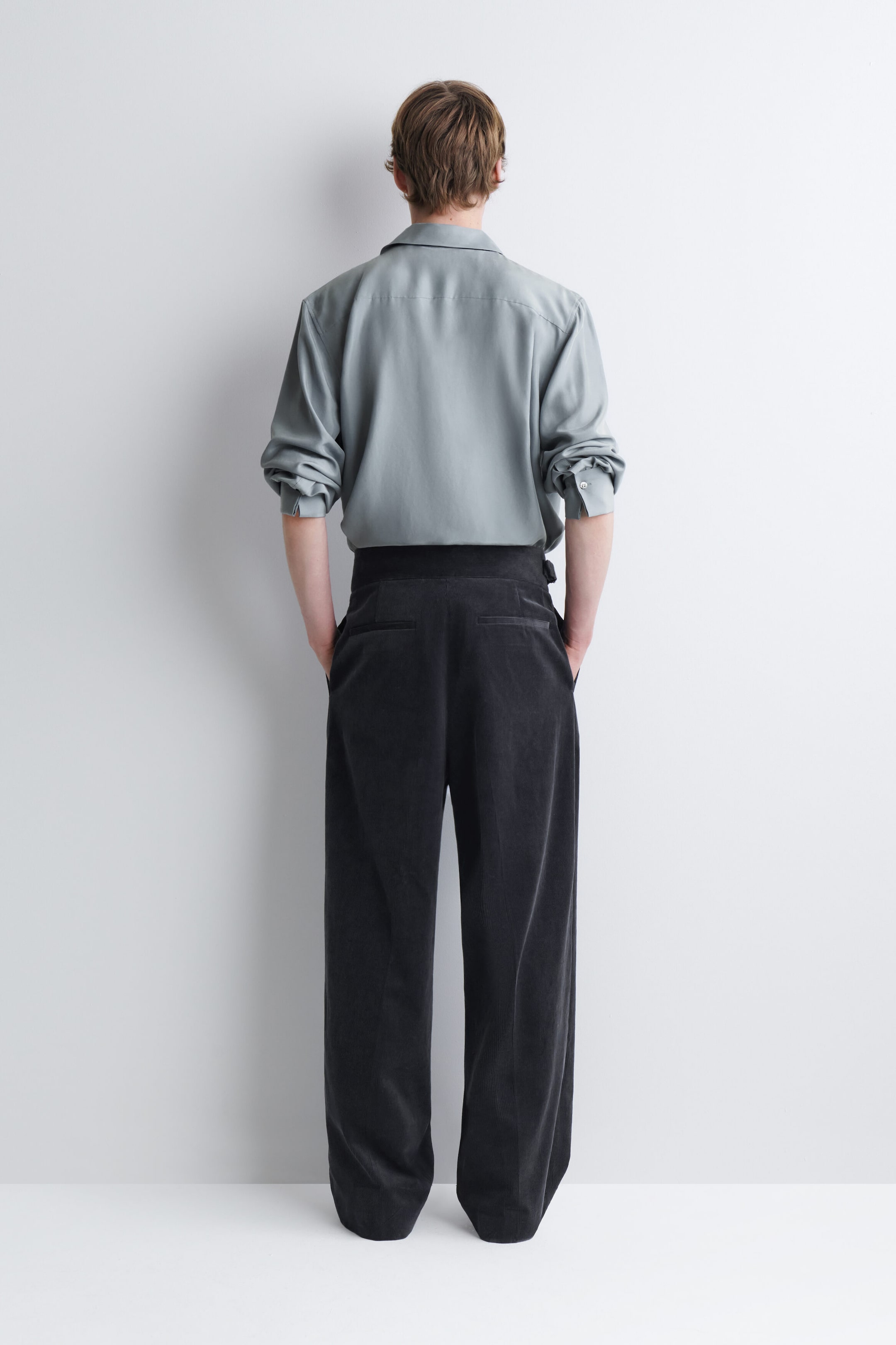These dark gray corduroy relaxed-fit wide-leg pants are worn by the model, who is standing with their back to the camera and hands in their pockets. The high-waisted pants feature rear welt pockets and are styled with a tucked-in light gray long-sleeve shirt. These dark gray corduroy relaxed-fit wide-leg pants are worn by the model, who is standing with their back to the camera and hands in their pockets. The high-waisted pants feature rear welt pockets and are styled with a tucked-in light gray long-sleeve shirt.