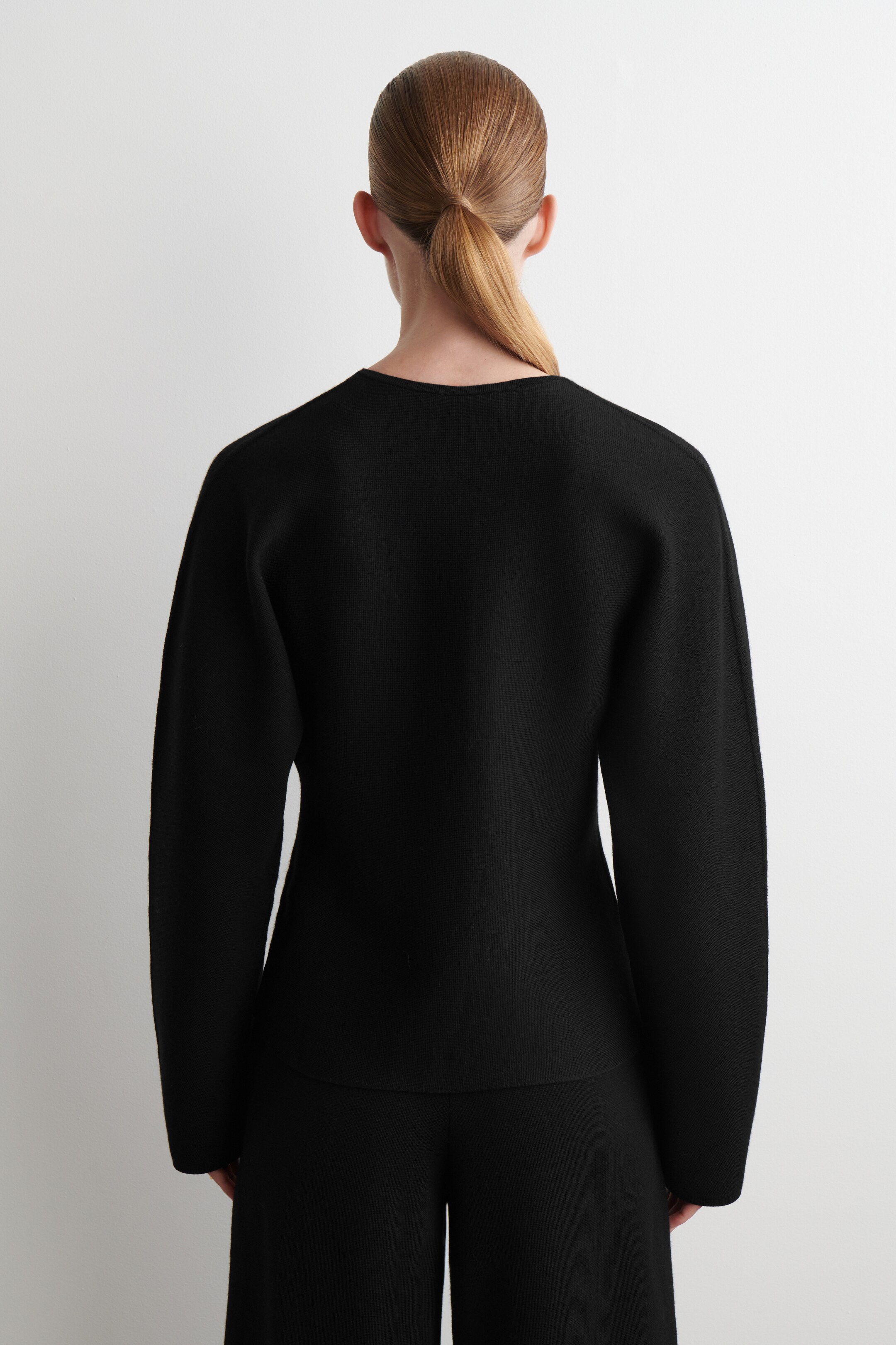 Größeres Bild ansehen: Ein schwarzes langärmliges Oberteil und passende weite Hosen, die aus einem Strickstoff zu bestehen scheinen, werden von hinten getragen. Das Oberteil zeichnet sich durch eine lockere Passform mit einem breiten, flachen Ausschnitt und fließenden langen Ärmeln aus, während die Hosen eine weite Silhouette aufweisen.