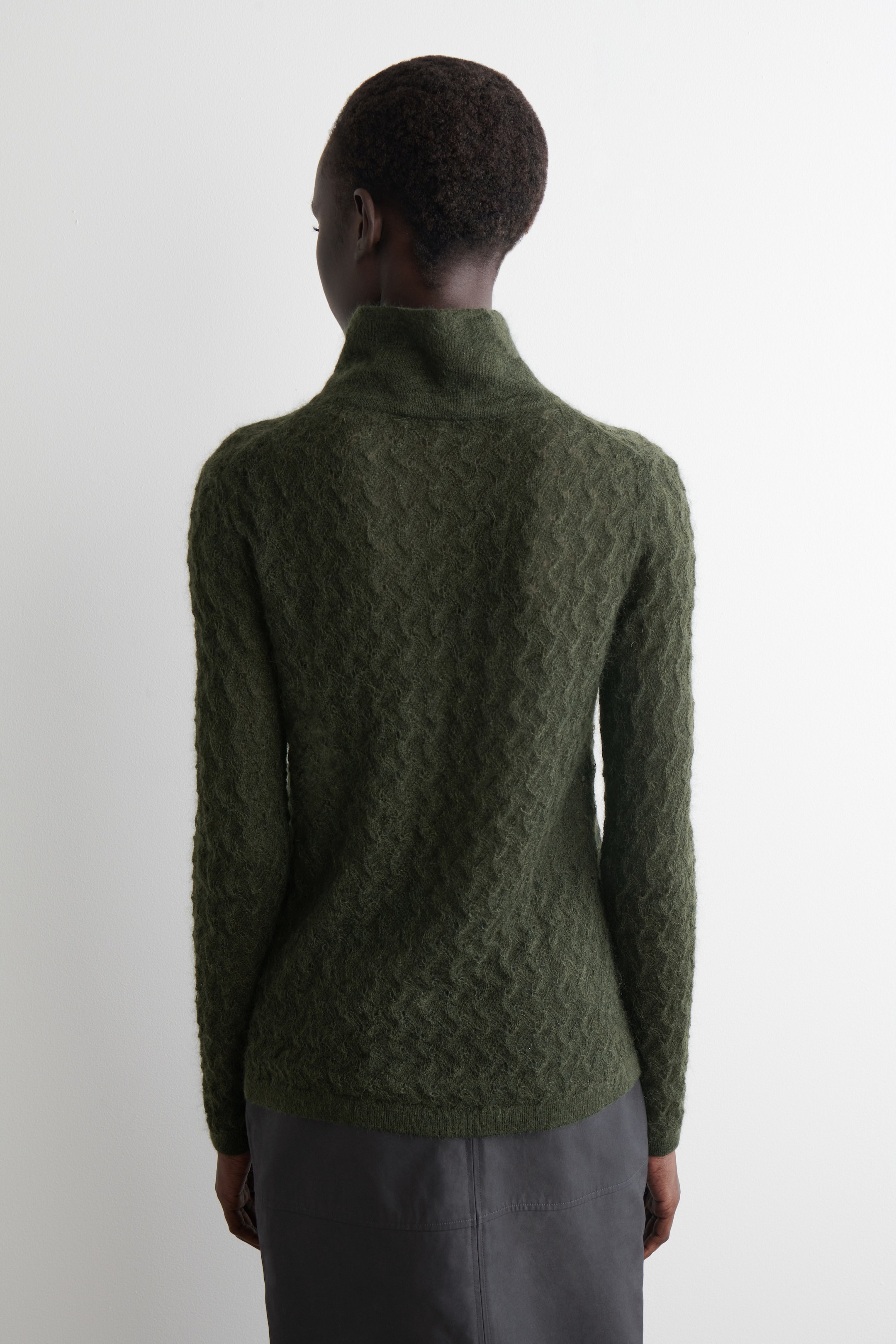Grösseres Bild ansehen: Ein dunkelgrüner Pullover wird von hinten betrachtet und zeigt ein wellenförmiges Strickmuster. Der Pullover hat einen Stehkragen und lange Ärmel, und sein Saum endet knapp über einem dunkelgrauen Rock.