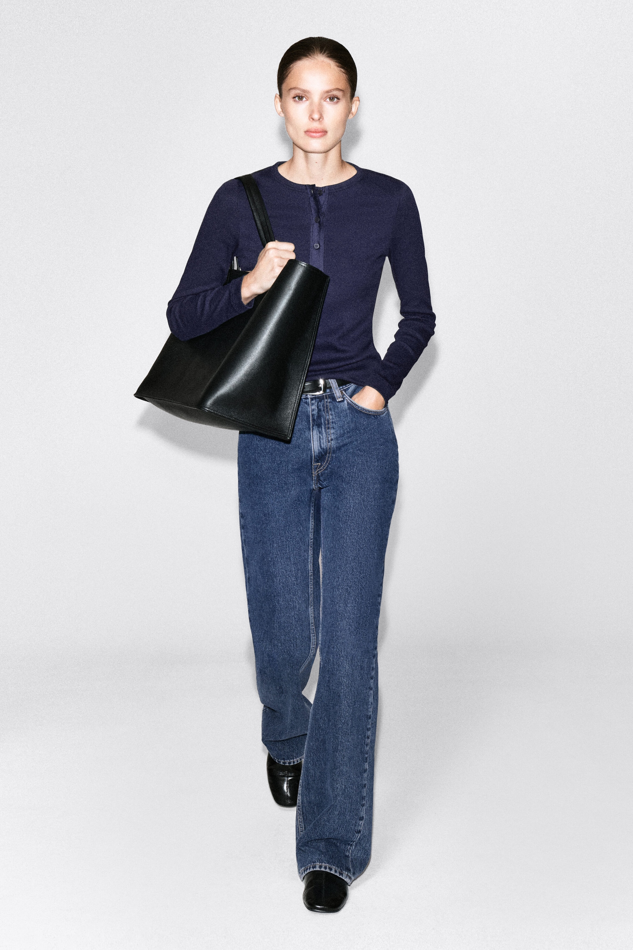 A ribbed cotton henley top in navy blue is being worn by the model, featuring a button placket and long sleeves. The top is tucked into blue jeans and paired with a large black leather tote bag carried over the shoulder. A ribbed cotton henley top in navy blue is being worn by the model, featuring a button placket and long sleeves. The top is tucked into blue jeans and paired with a large black leather tote bag carried over the shoulder.