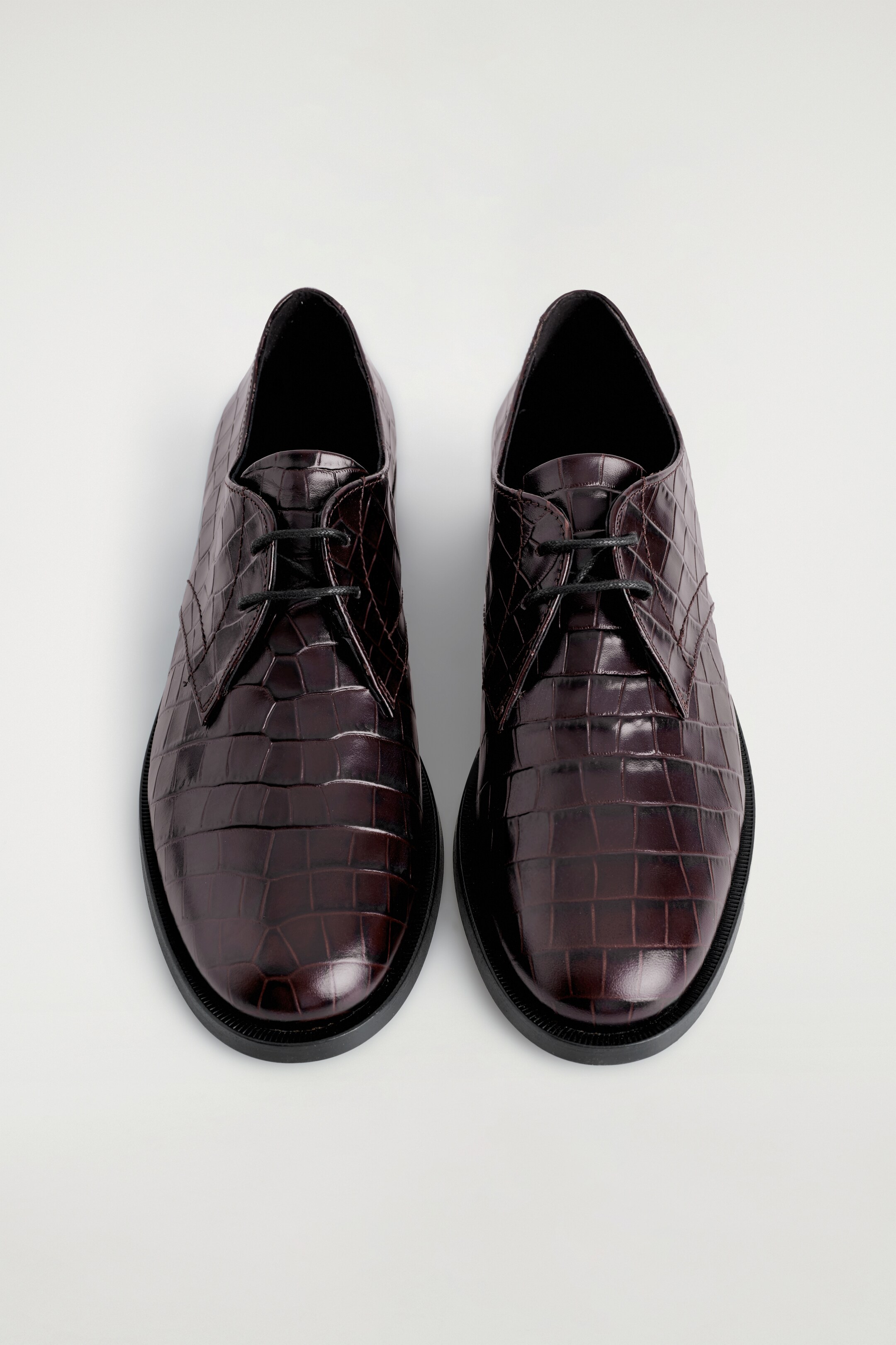 Visualizza immagine più grande: Due paia di scarpe Derby color espresso sono affiancate, caratterizzate da una tomaia in pelle con stampa coccodrillo, punte arrotondate e lacci in cotone cerato nero. Una suola nera è visibile intorno alla base di ciascuna scarpa.
