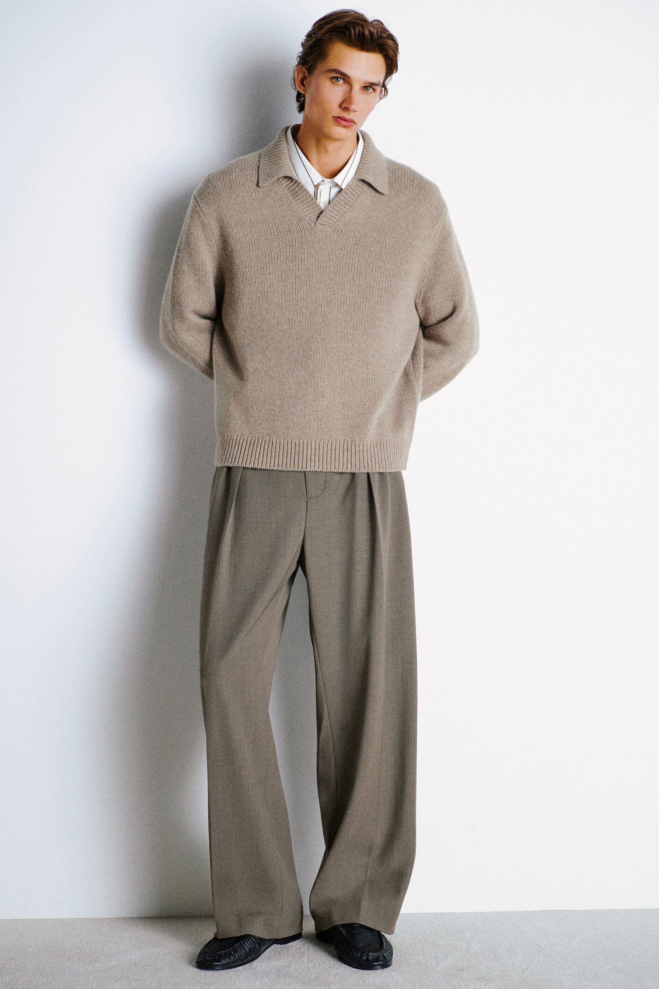 A model is wearing a beige, relaxed-fit cashmere polo sweater layered over a white collared shirt, paired with taupe wide-leg trousers, facing forward. The knit sweater features an open collar and a ribbed hem, while the trousers have deep front pleats and central pressed creases. A model is wearing a beige, relaxed-fit cashmere polo sweater layered over a white collared shirt, paired with taupe wide-leg trousers, facing forward. The knit sweater features an open collar and a ribbed hem, while the trousers have deep front pleats and central pressed creases.